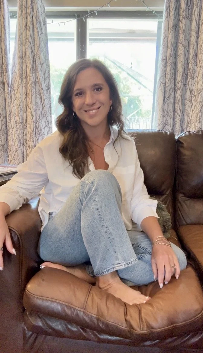 A woman with long wavy hair sitting cross-legged on a brown leather couch, smiling at the camera, in a room with large windows and patterned curtains.
