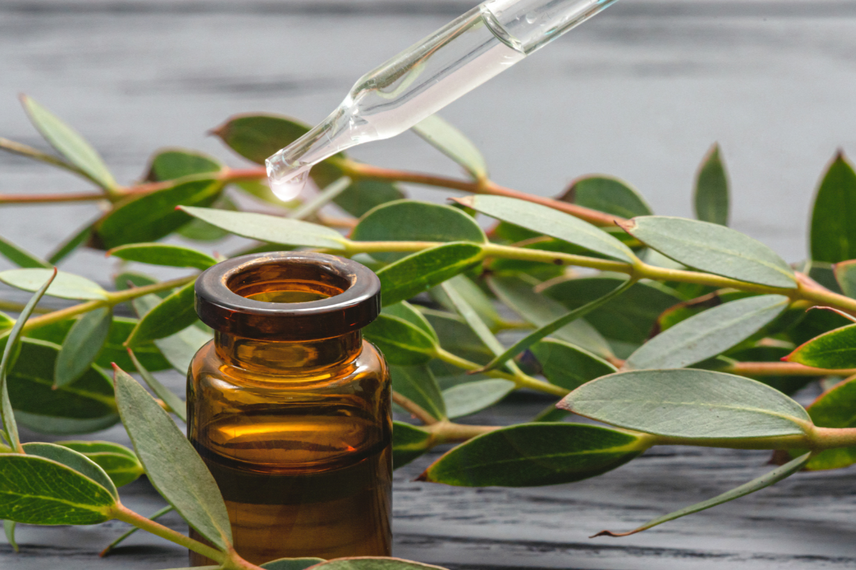 An amber glass dropper bottle with a glass pipette above it, set against green leaves on a gray wooden surface.