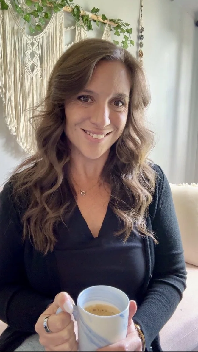 A woman with wavy brown hair smiling and holding a cup of coffee, sitting in a cozy, decorated space with macrame wall hangings and greenery in the background.
