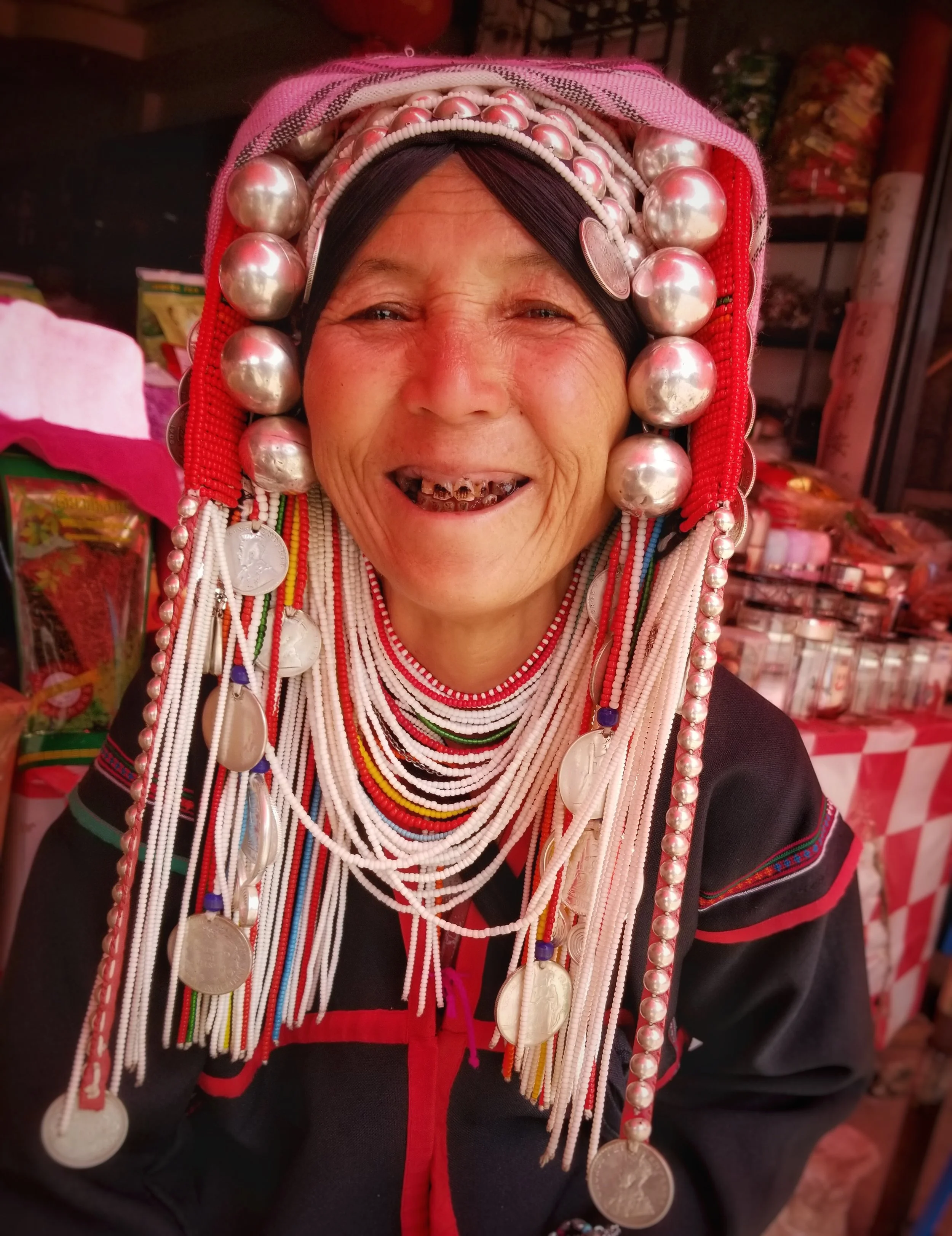 A smiling elderly woman wearing traditional attire with numerous colorful beaded necklaces and large silver coin jewelry. She is outside in a market setting with various items in the background.