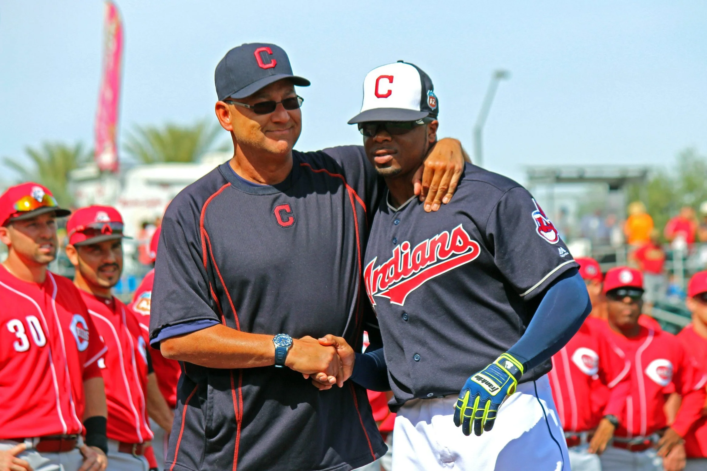 Two baseball players from the Cleveland Indians team shaking hands on the field, with other team members standing in the background.