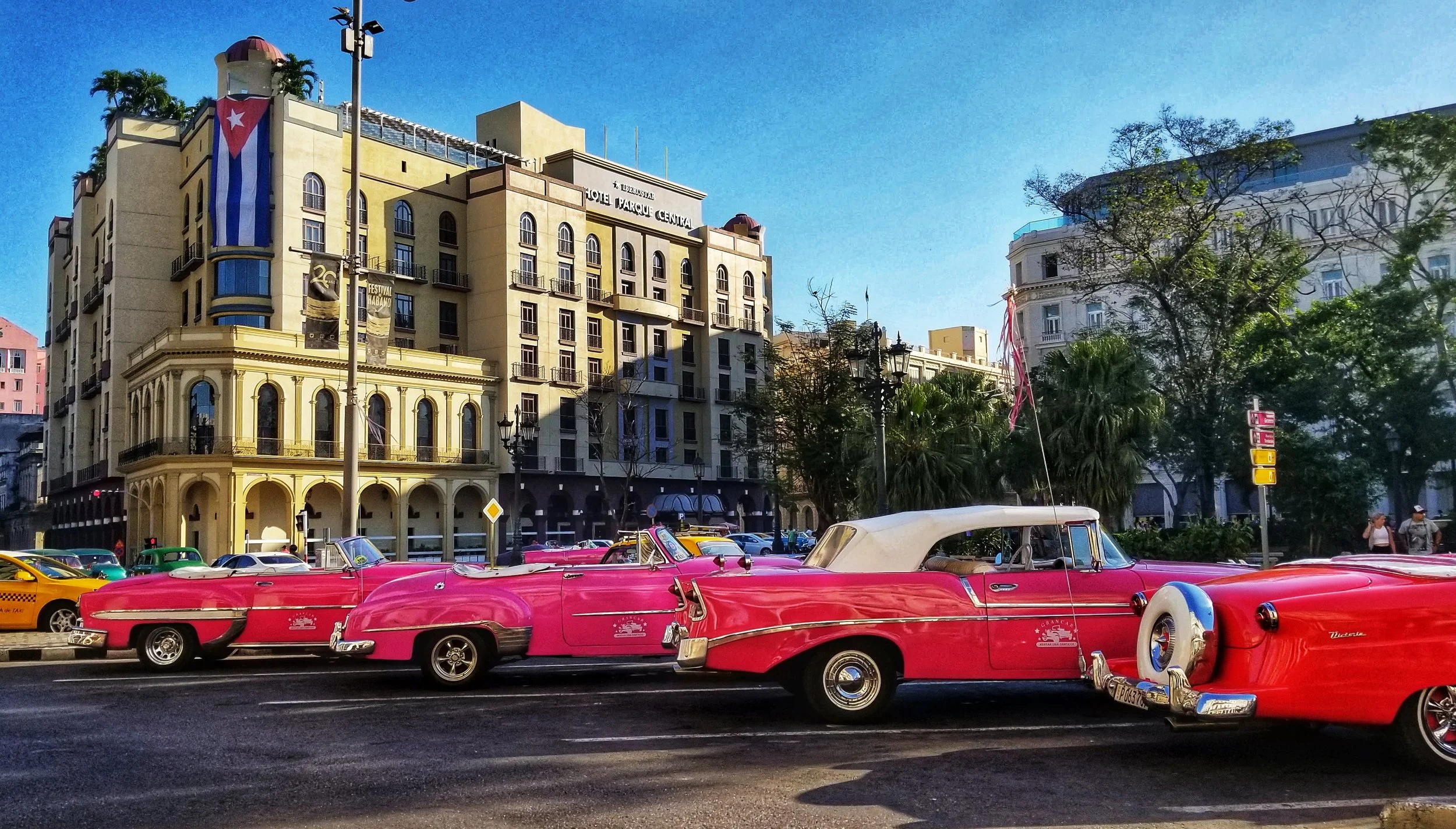 Street scene with pink classic cars parked in front of a yellow building with arched windows and balconies, Cuban flag hanging from a pole, and trees with a clear blue sky overhead.