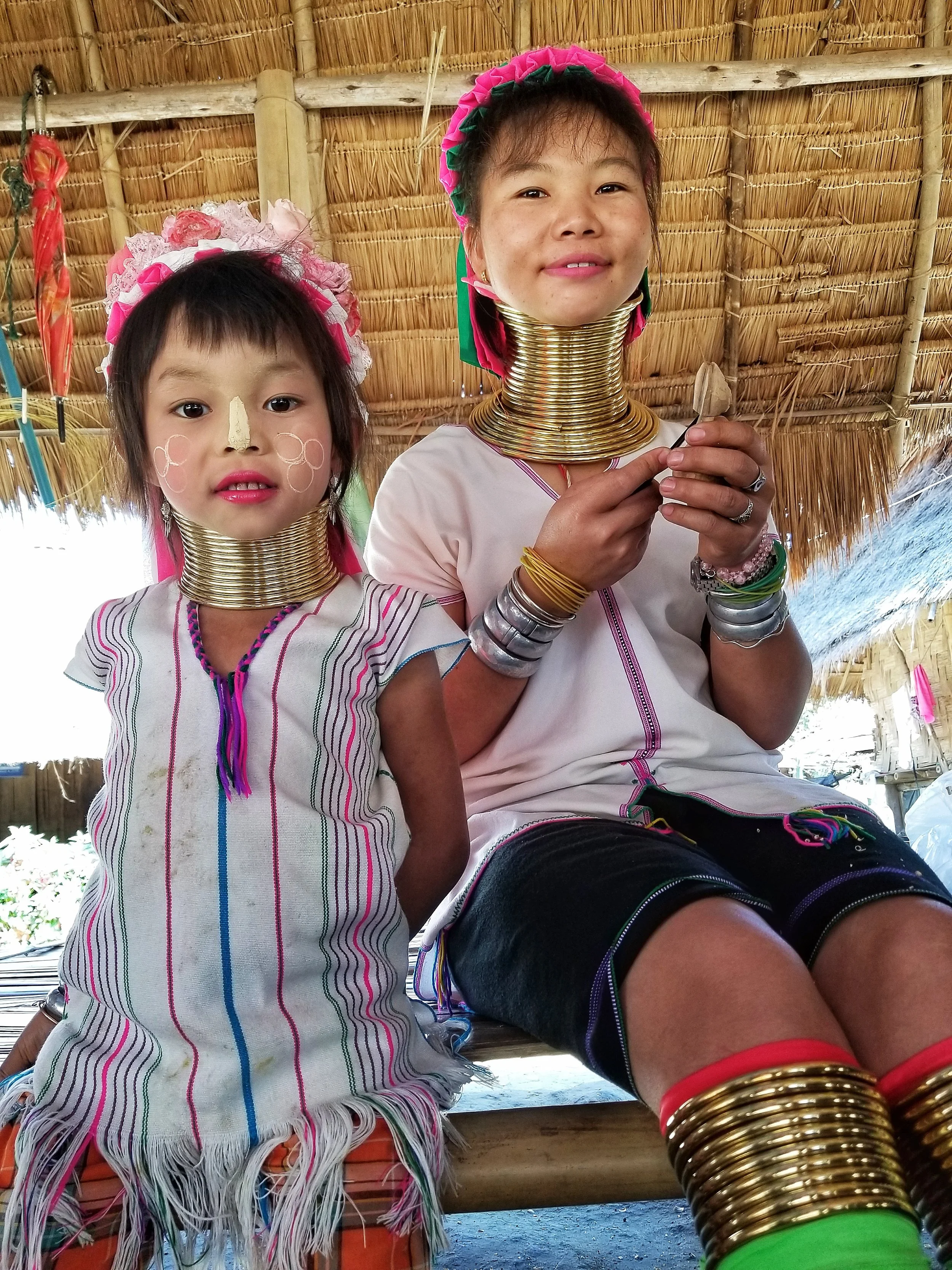 Two women, likely part of the Kayan Lahwi tribe, wearing traditional brass neck rings and colorful clothing, sitting under a thatched roof, with one woman smiling at the camera and the other looking directly at it.