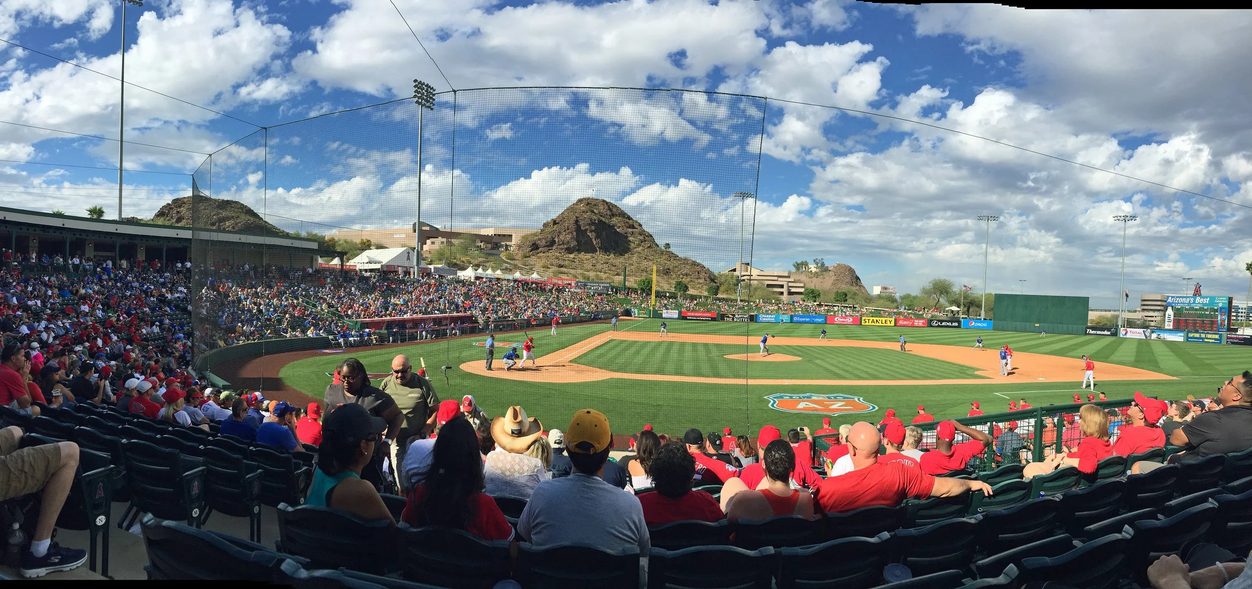 Crowd at a baseball game in a stadium with Arizona mountains in the background, players on the field, and blue sky with clouds overhead.