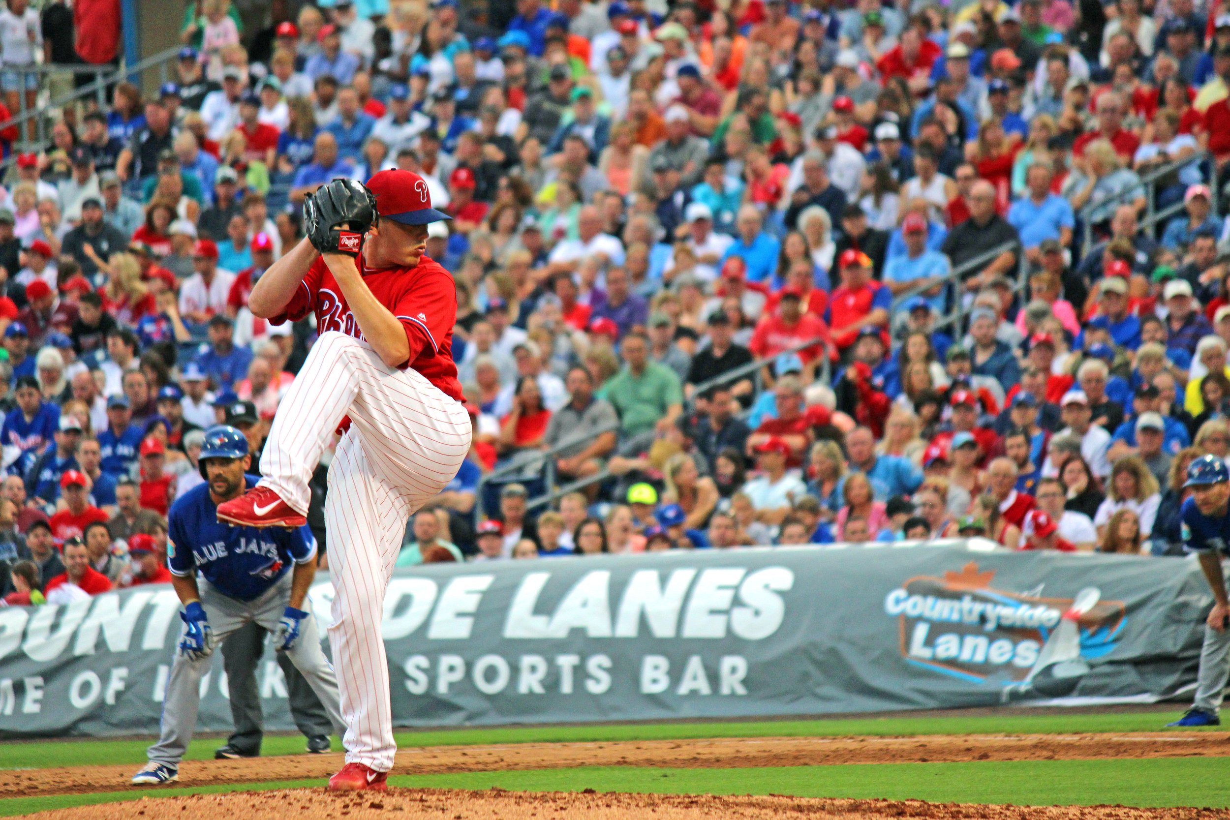 A baseball pitcher in a red jersey and cap preparing to throw a pitch during a game. The stands are filled with spectators.