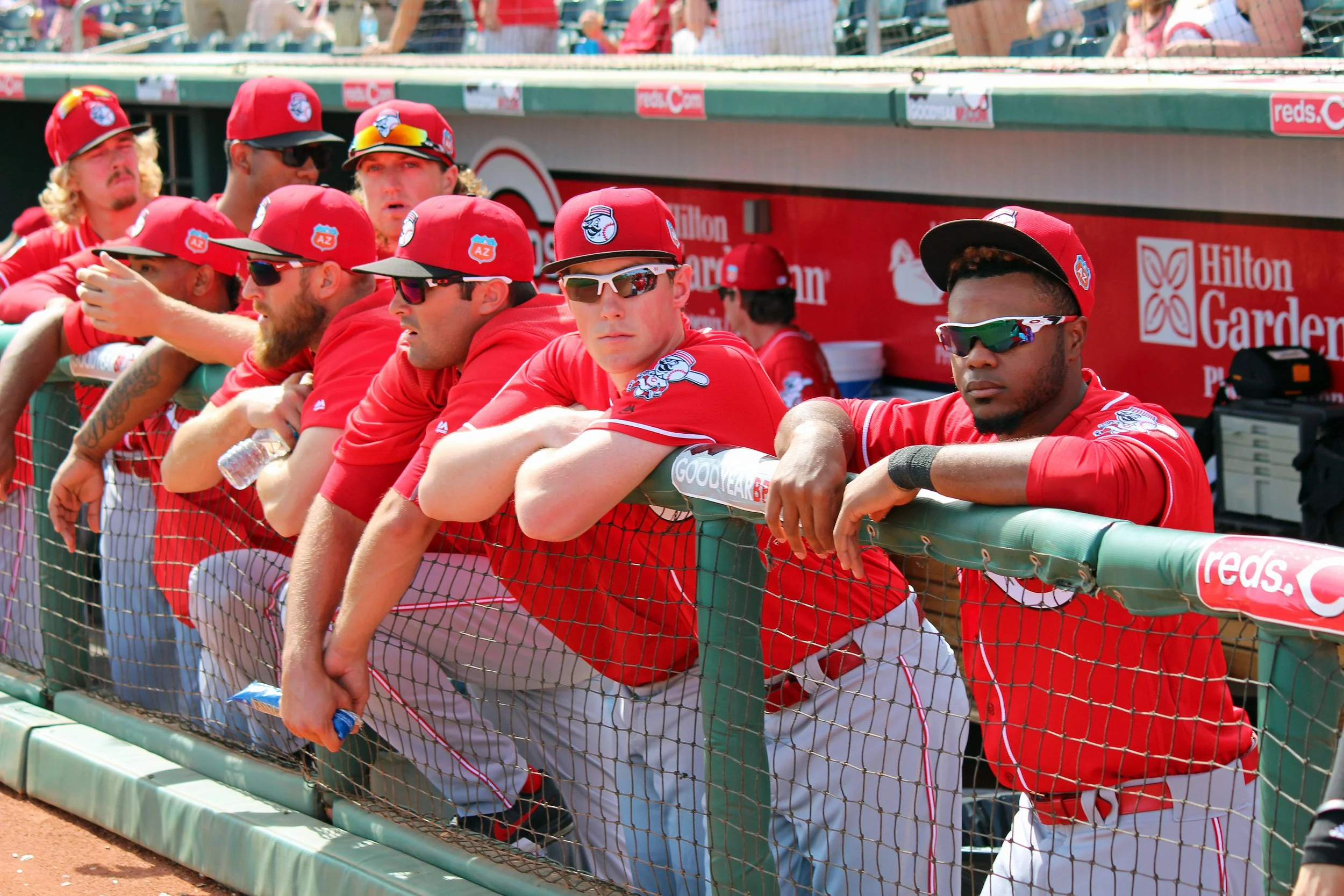 Baseball players in red uniforms and caps sitting and standing along the dugout fence during a game.