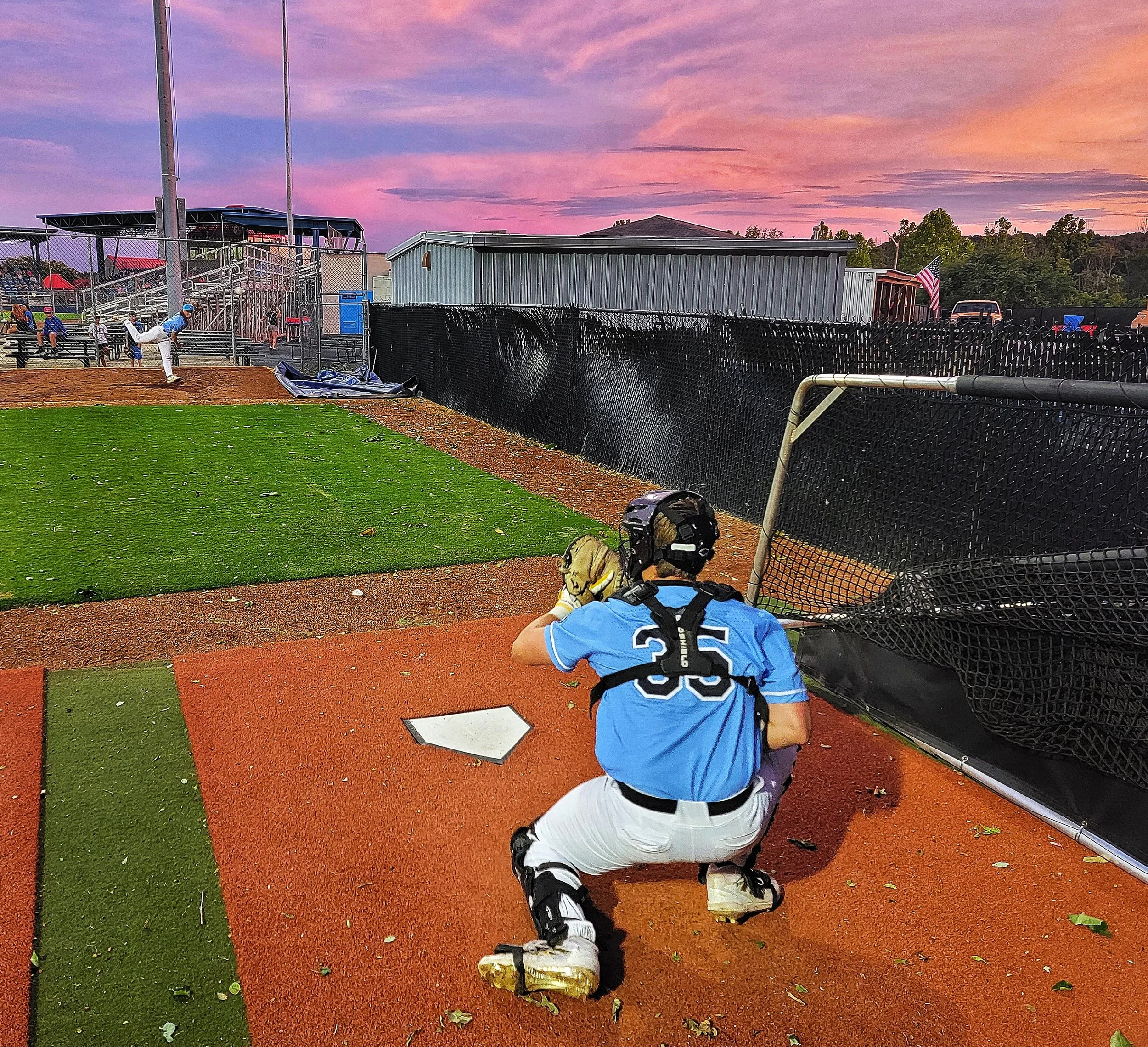 A young baseball player with golden hair kneeling on the ground near a baseball field fence, looking at a fallen balloon. The scene is set against a colorful sunset sky with pink, purple, and orange hues. The player is wearing a blue and white unifor