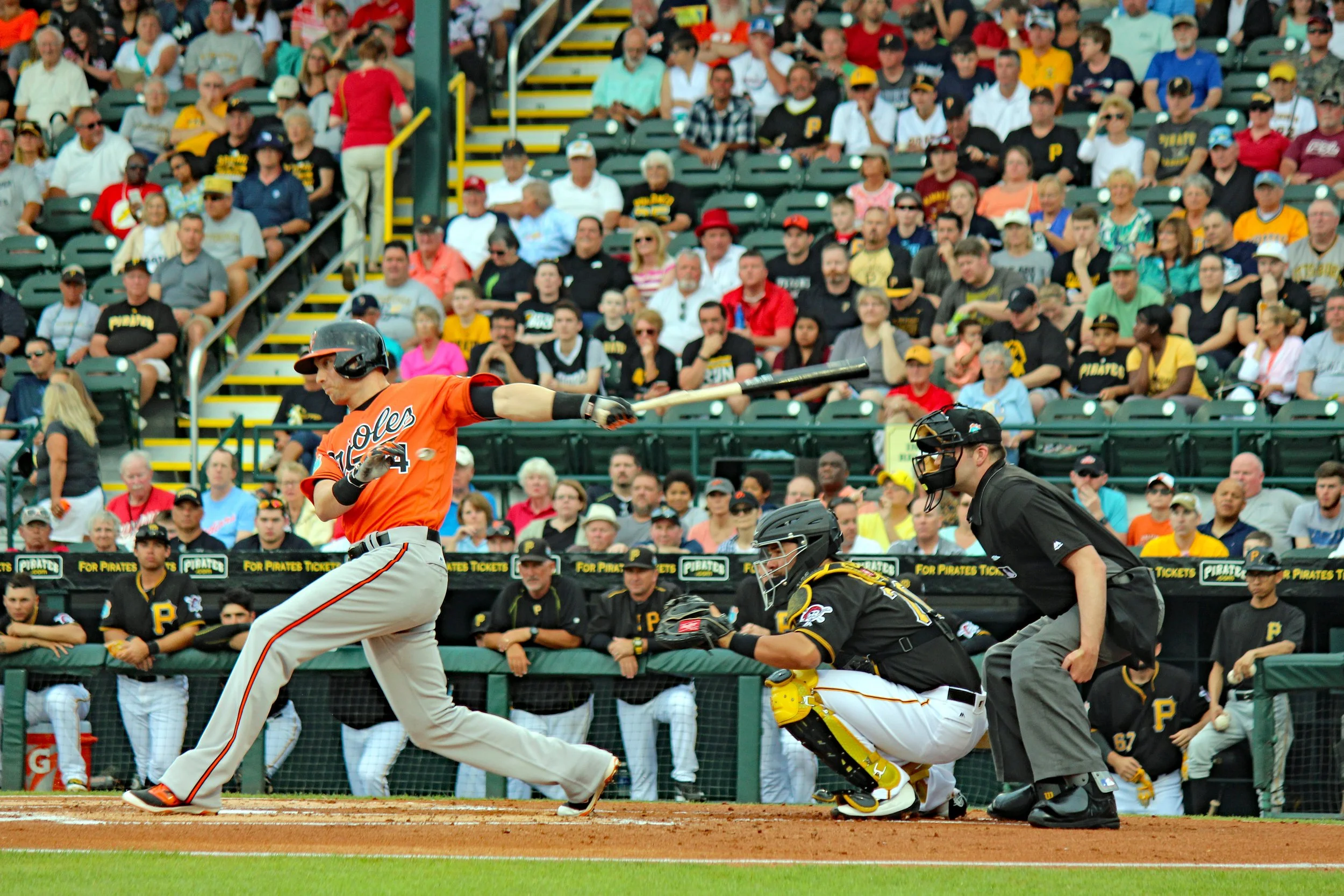 A baseball player in an orange uniform swinging a bat at a pitch during a game. The catcher or umpire is kneeling behind home plate, and there is a crowd of spectators in the background.