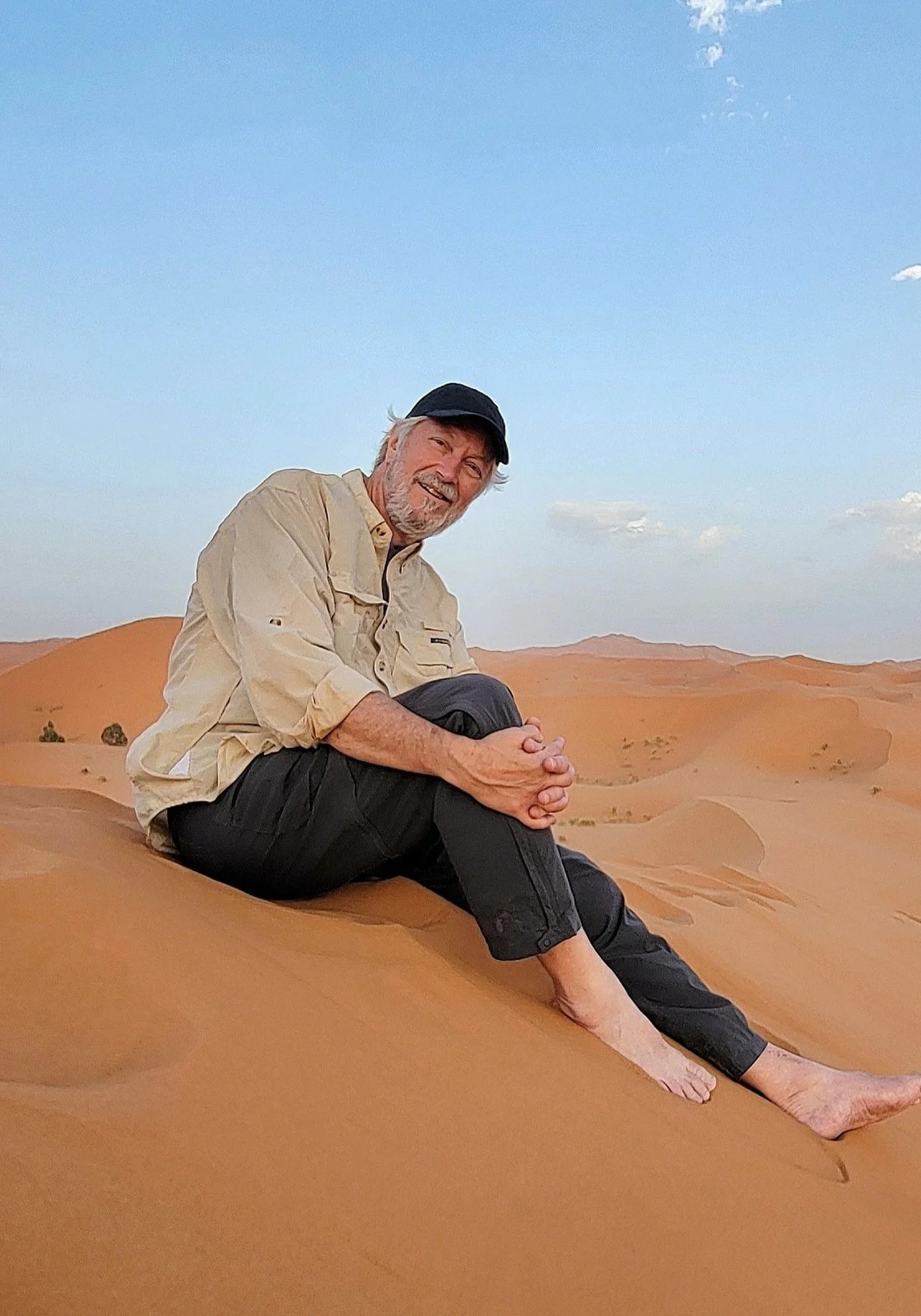 An older man with gray hair and a beard sitting on sand dunes in a desert landscape, smiling, wearing a beige shirt and black pants, with a black cap, with a clear blue sky and scattered clouds in the background.