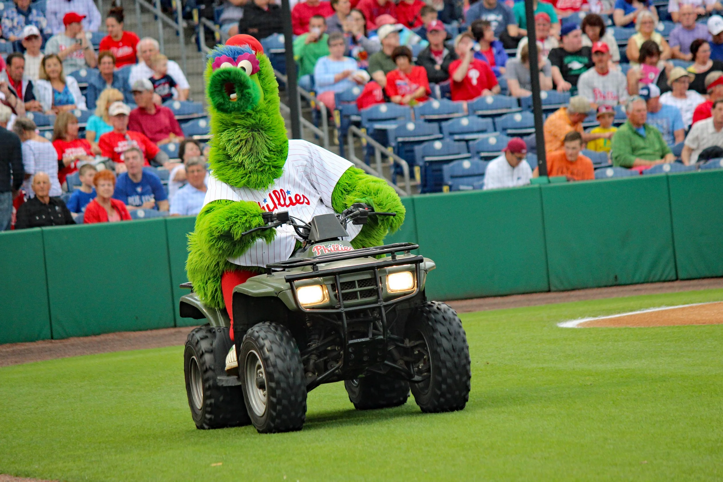 A green mascot with a furry face wearing a baseball jersey riding a small ATV on a baseball field during a game with spectators in the stands.