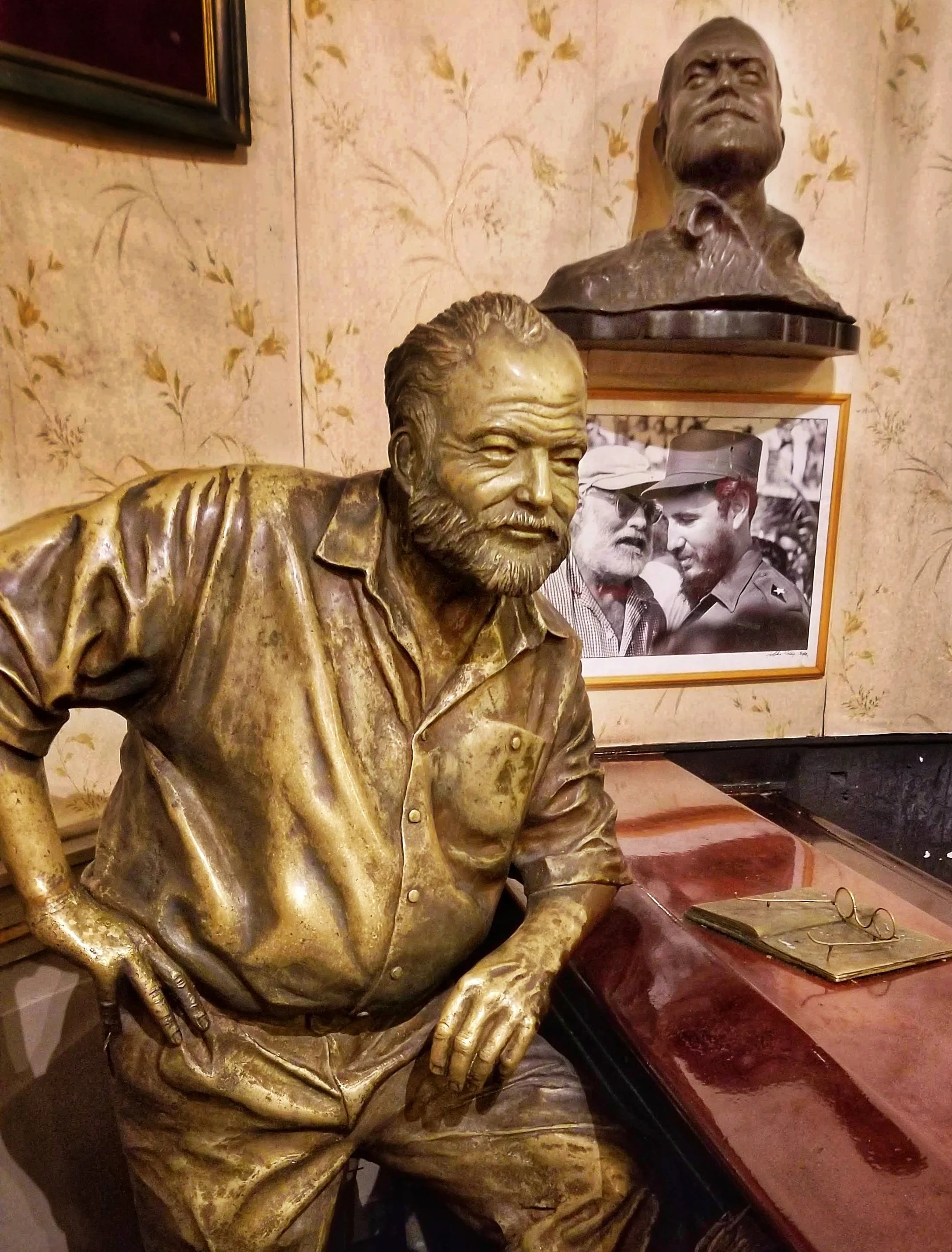 A bronze bust of a man with a beard and mustache, dressed in a collared shirt and jacket, sitting at a wooden table with glasses, a framed photograph in the background, and a pair of glasses resting on the table.