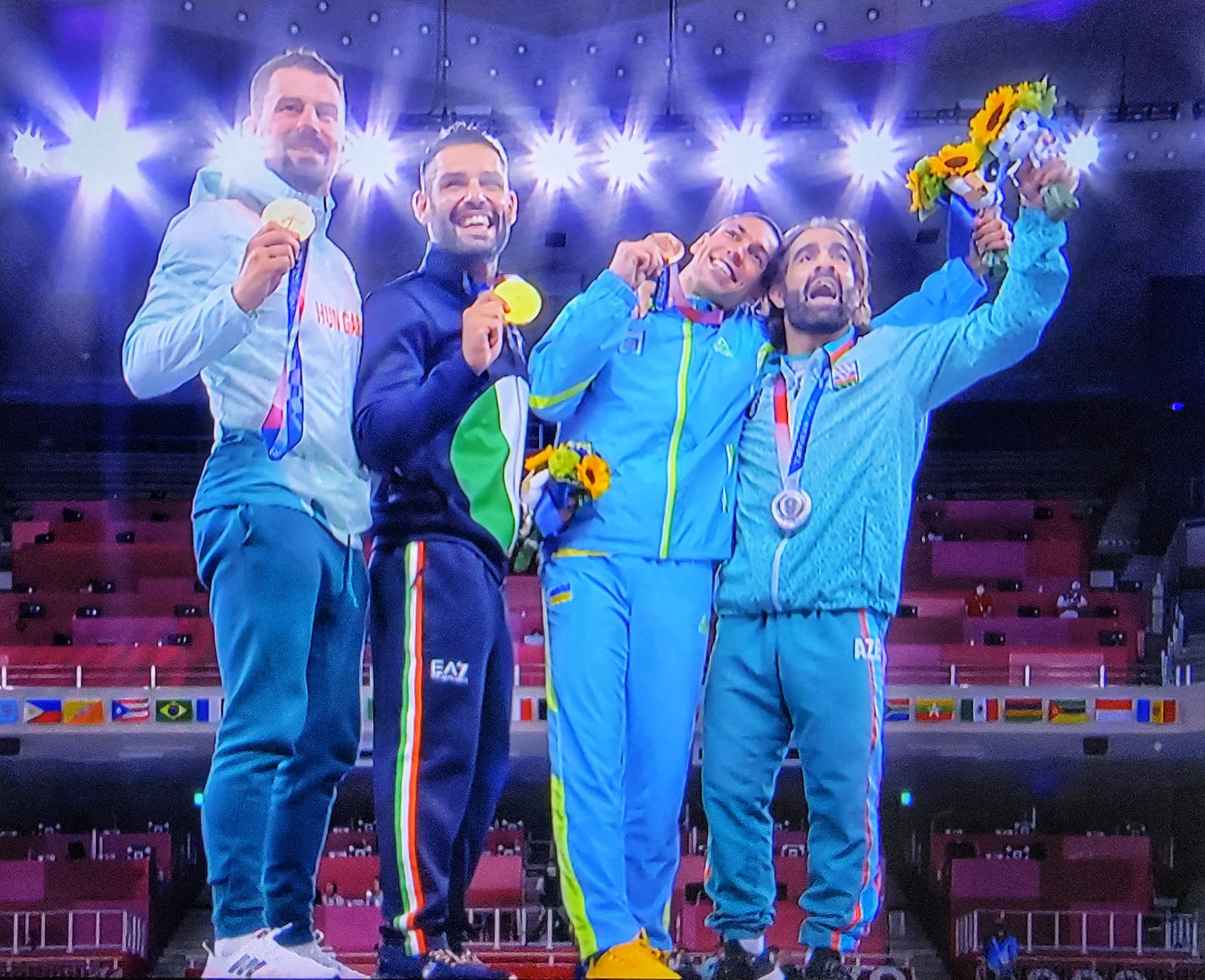 A group of five athletes on a podium holding medals, celebrating their victory. They are wearing sports outfits with an Olympic or World Championship theme, one holding a bouquet of sunflowers, and the background features bright lights and a display 