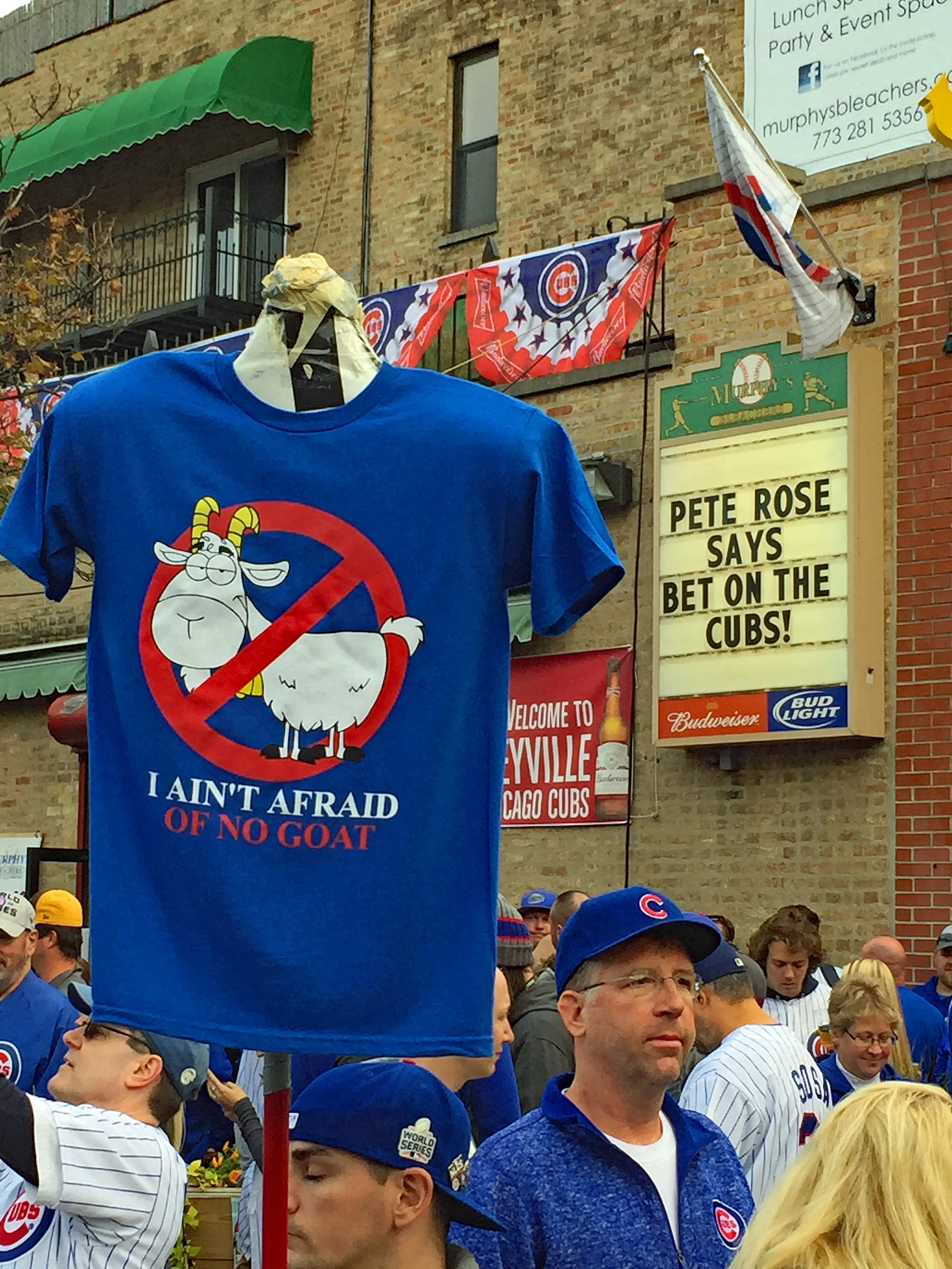 A crowd of Chicago Cubs fans gathered outdoors during daytime, with some wearing Cubs gear and hats. A blue T-shirt with a cartoon goat and the text ‘I AIN’T AFRAID OF NO GOAT’ is prominently displayed. In the background, signs and banners related to