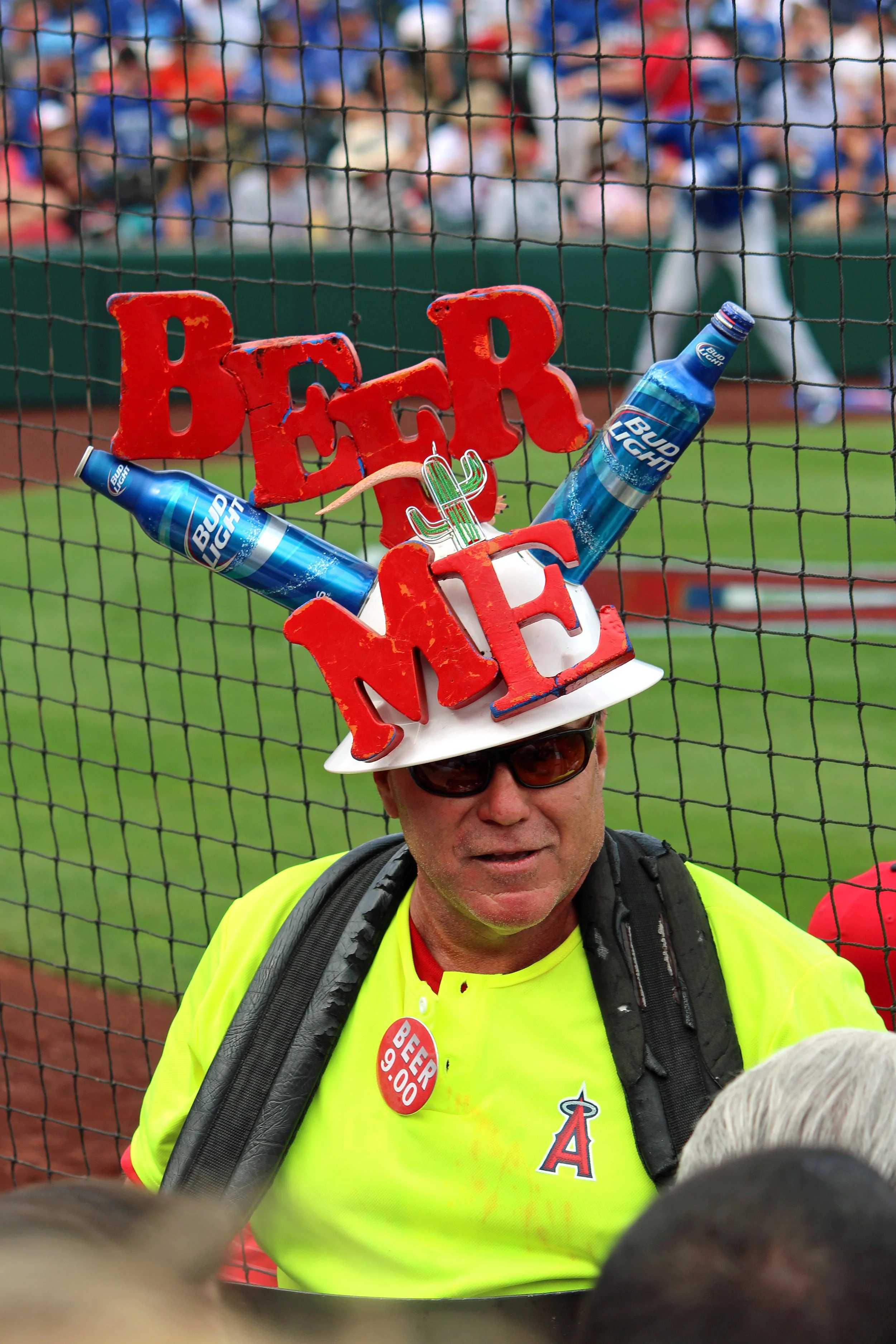 A man wearing sunglasses and a yellow Angels baseball jersey with a backpack, standing in front of a dugout fence at a baseball game. He sports a colorful hat arrangement spelling 'BEER,' featuring two Bud Light bottles attached to it.