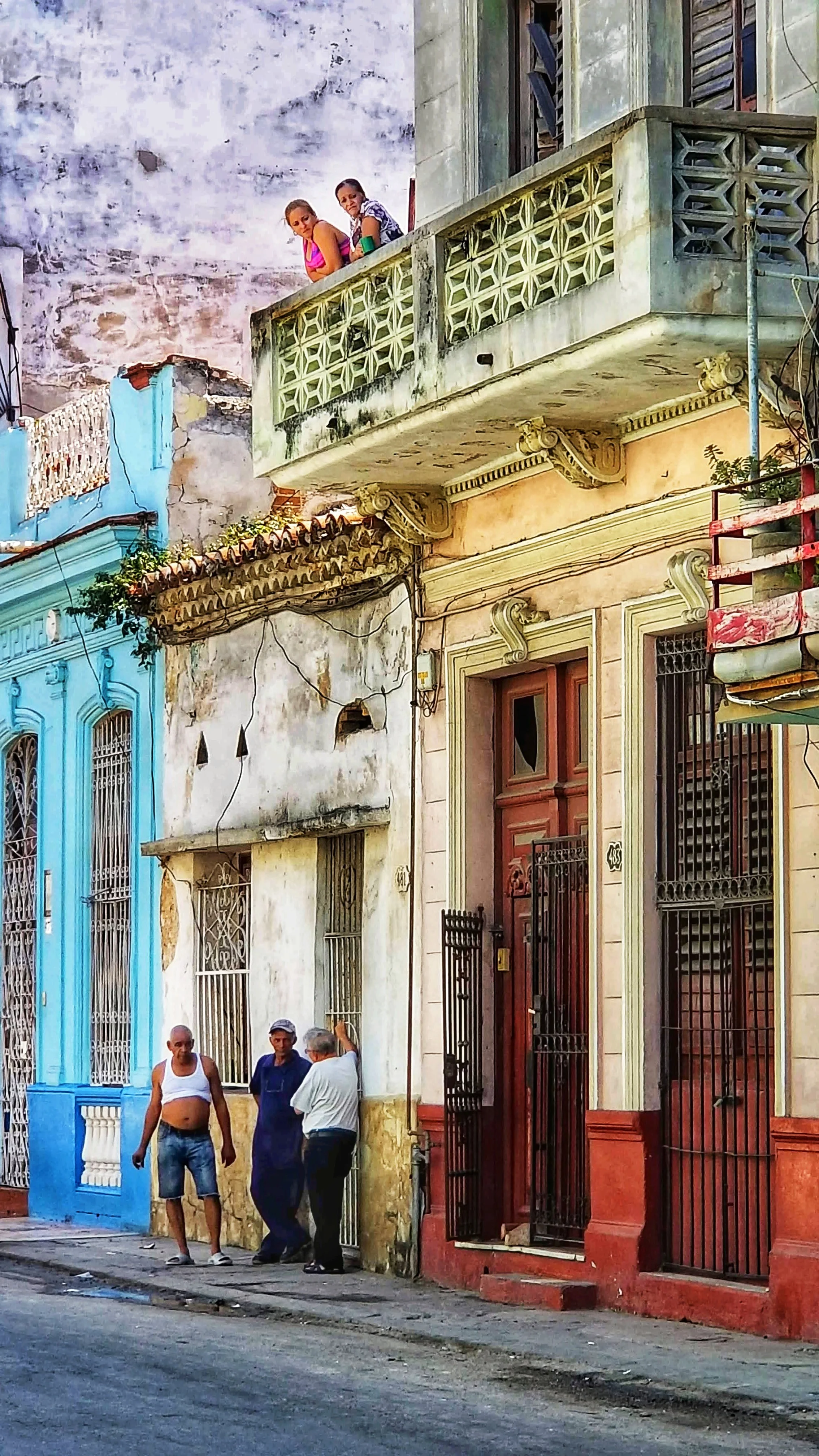 Street scene with colorful old buildings and four men standing on sidewalk, two of them talking and one shirtless.
