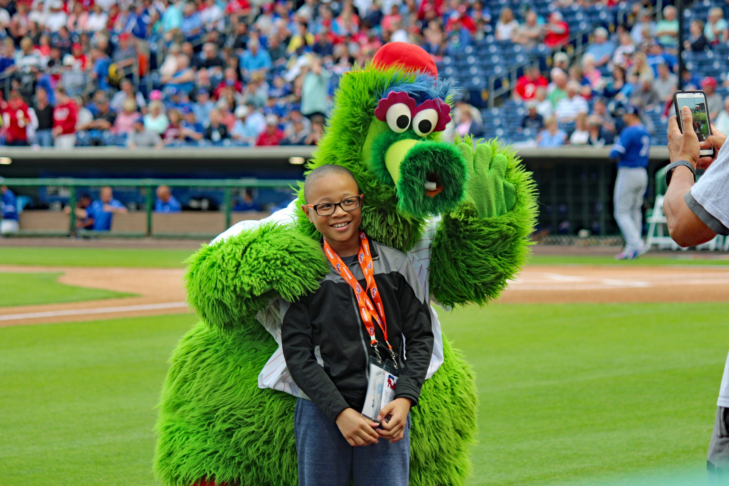 A child with glasses standing next to a person in a large green furry mascot costume with googly eyes and a red tuft on its head on a baseball field, with spectators watching from the stands.