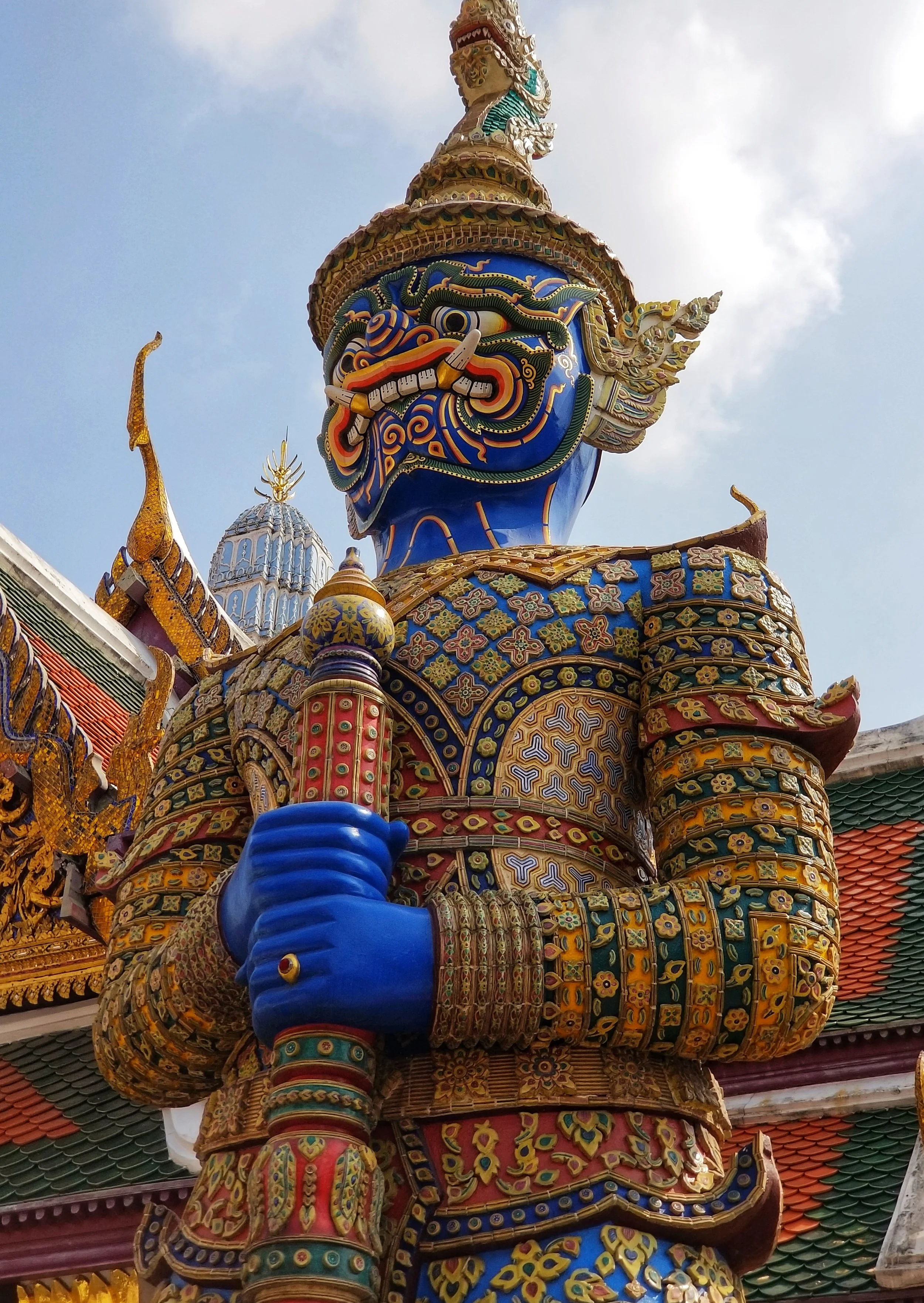 A large, ornate statue of a mythical guardian figure with a vibrant blue face, fierce expression, sharp teeth, and elaborate decorations, holding a weapon. The statue is decorated with intricate gold and colorful patterns, with a temple roof visible 