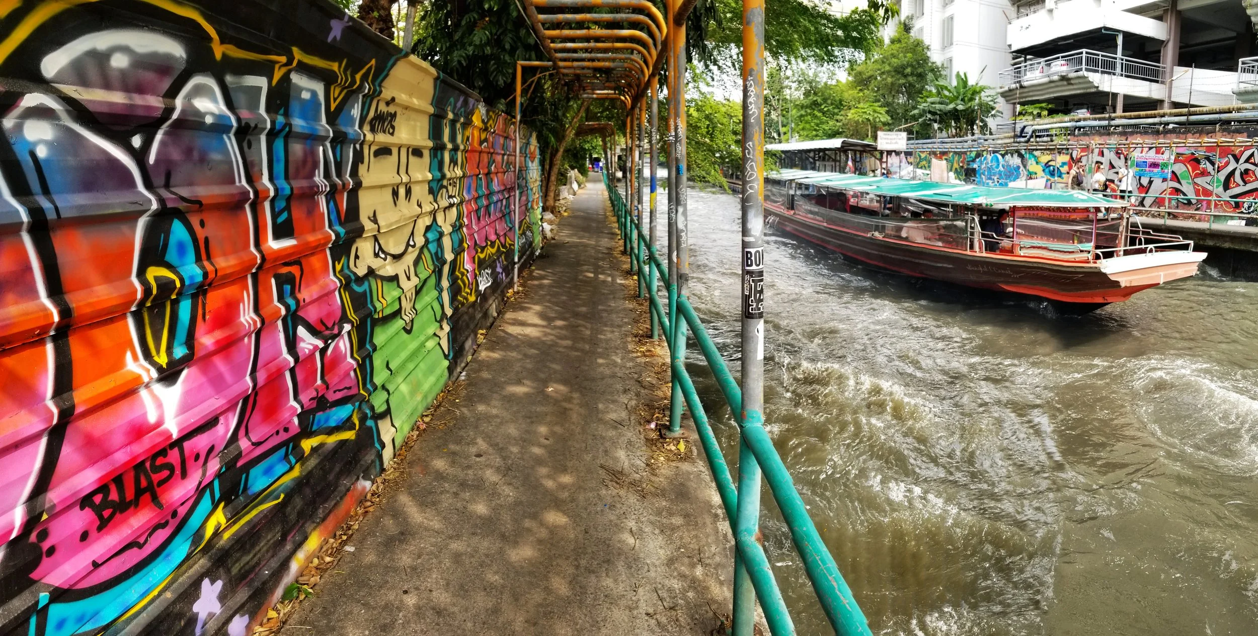 A river scene with a boat on the water near a colorful graffiti-covered wall and a sidewalk with a metal railing. Trees and buildings are visible in the background.