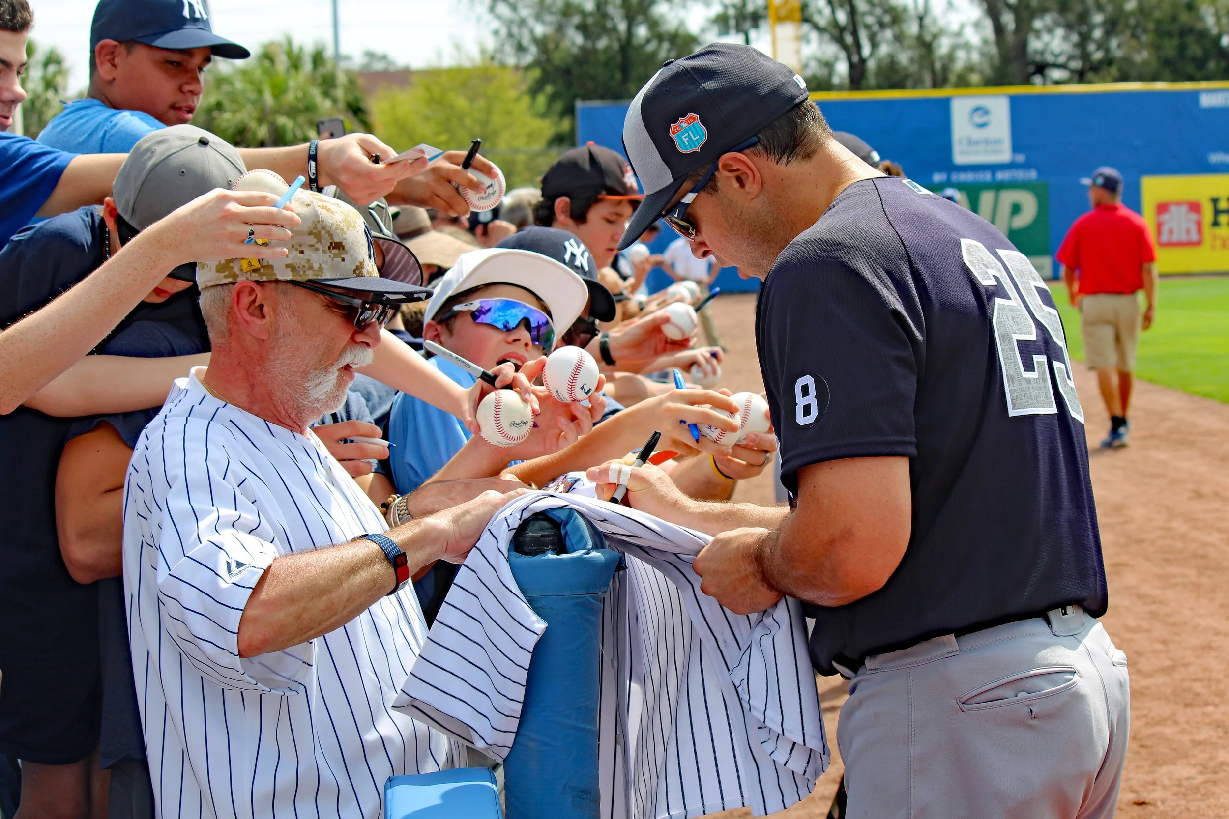 A baseball player signing autographs for young fans behind a fence at a baseball field, with fans holding baseballs and wearing caps, while a man in a red shirt stands in the background.