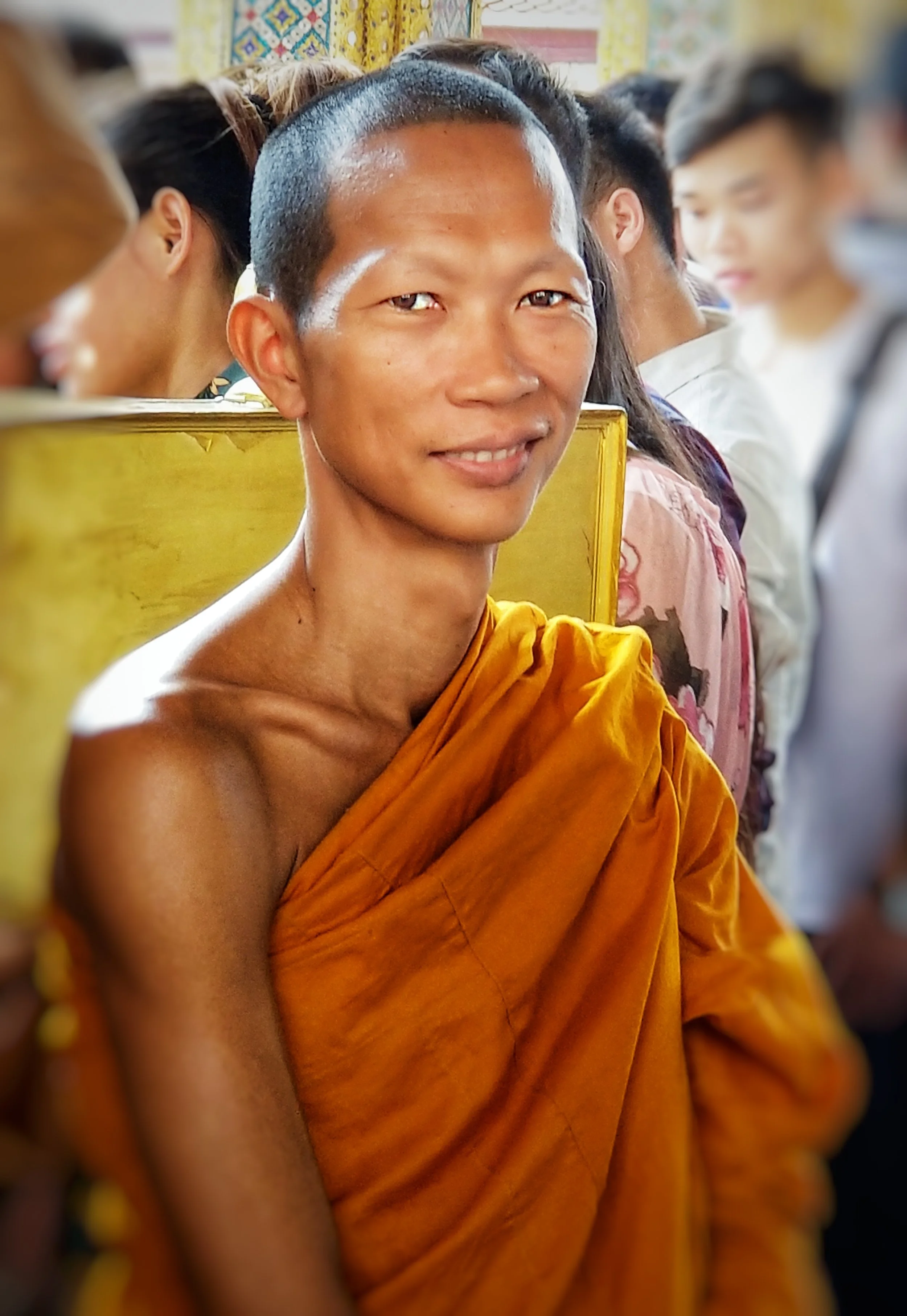A smiling Buddhist monk wearing orange robes sits in a line with others in brightly decorated surroundings.