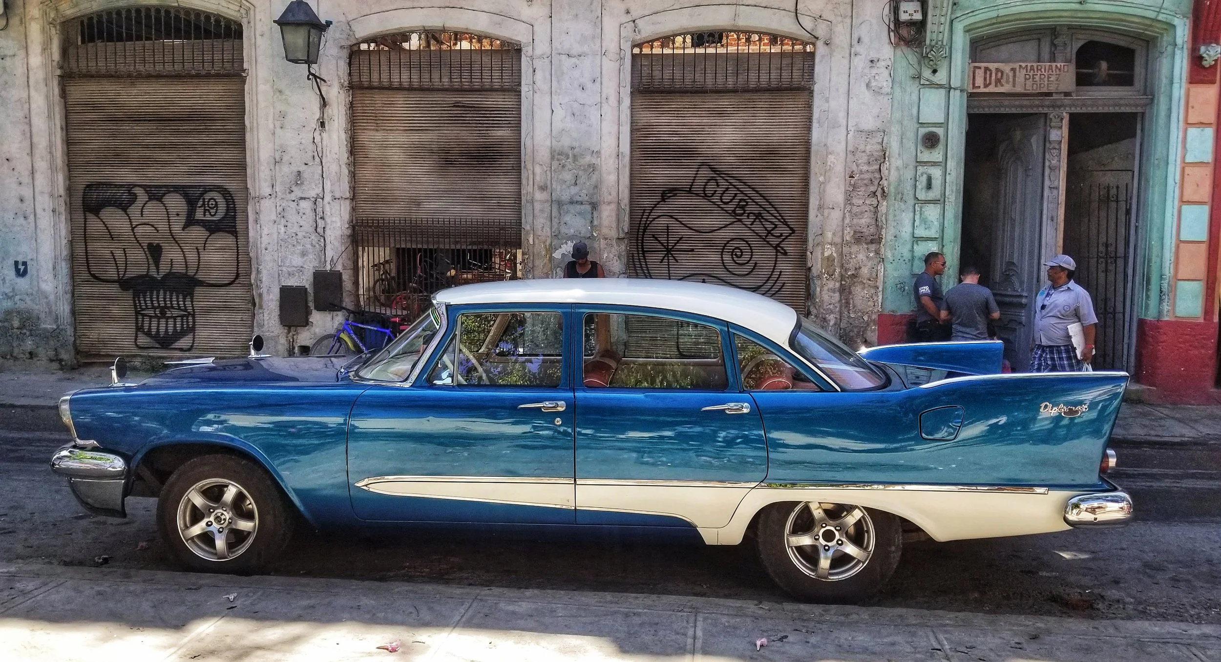 A vintage blue and white classic car parked on the street in front of an old building with graffiti-covered metal shutters and a colorful facade. Three men are standing near the building, engaged in conversation.