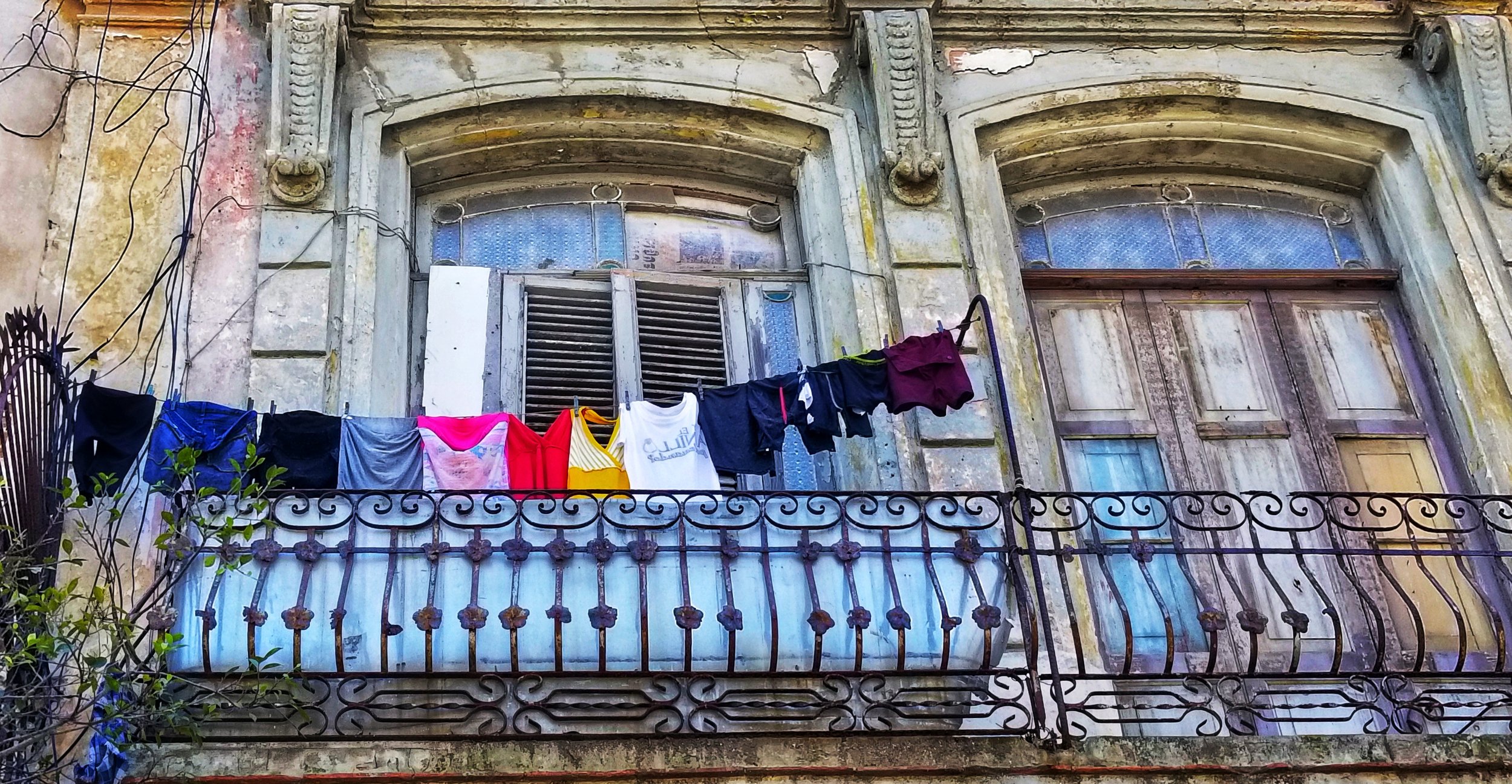Old building with worn paint, a small balcony with hanging laundry including shirts and shorts, and a rusted decorative metal railing in front.