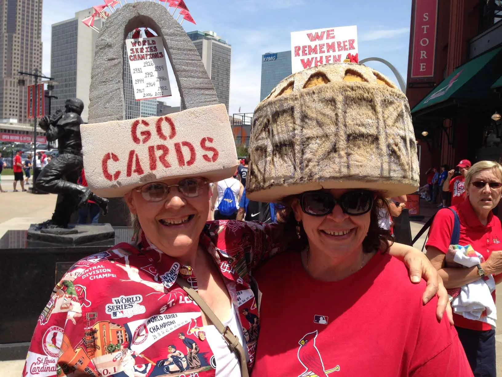 Two women at a sports celebration with carved bread hats, one reading 'GO CARDS' and the other with 'WE REMEMBER WHEN,' and a background of city buildings and a statue.