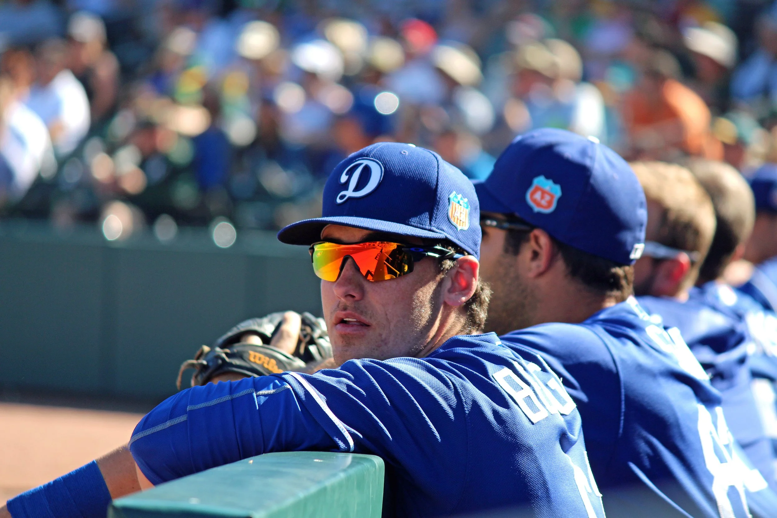 A baseball player wearing a blue cap and uniform sitting on the sidelines, with other players and spectators blurred in the background.