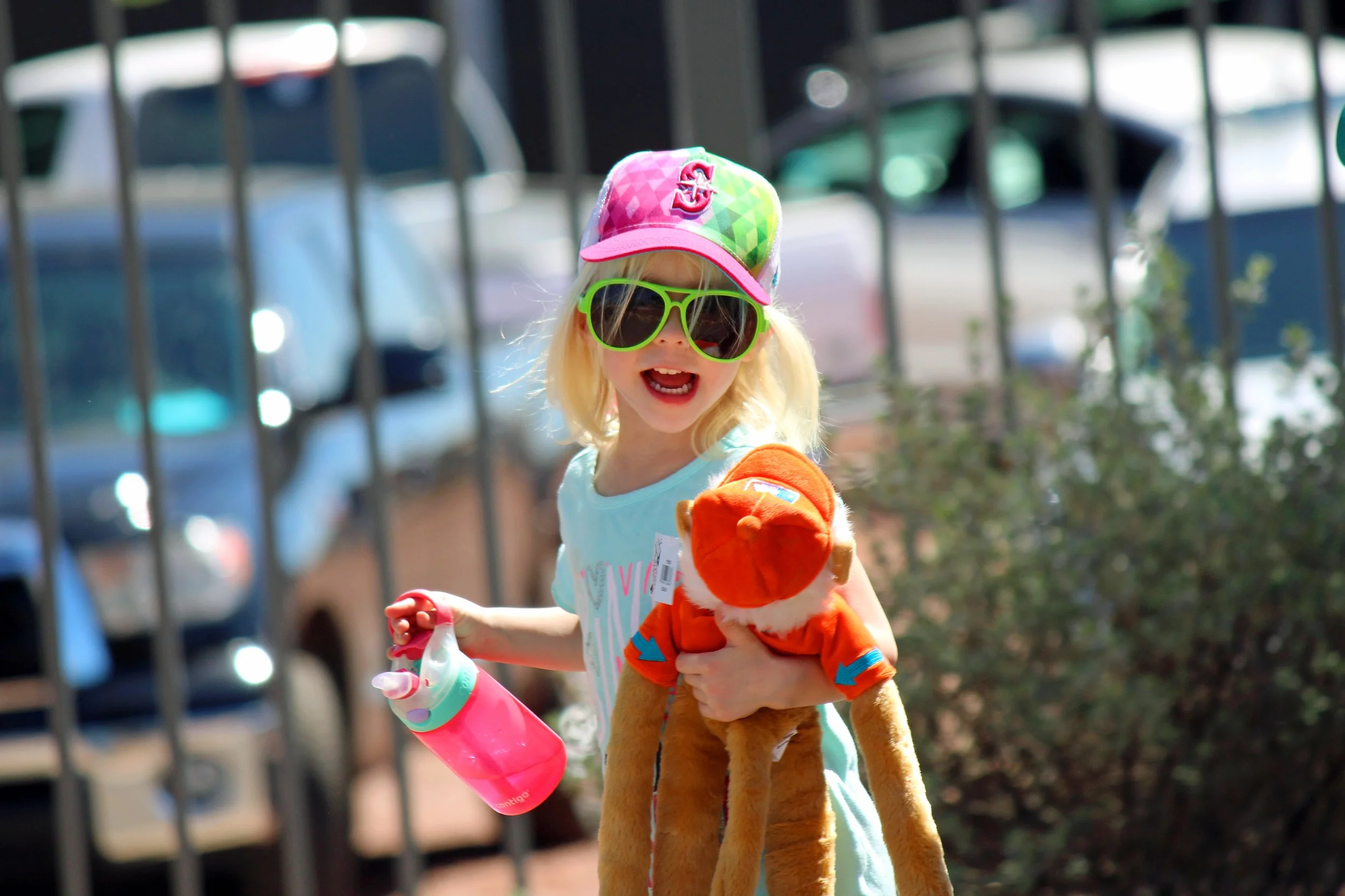 A young girl with blonde hair wearing large green sunglasses, a colorful baseball cap, and a light blue shirt. She is holding a pink water bottle and a plush tiger toy. The girl appears to be outdoors, near a metal fence, with cars and greenery in th