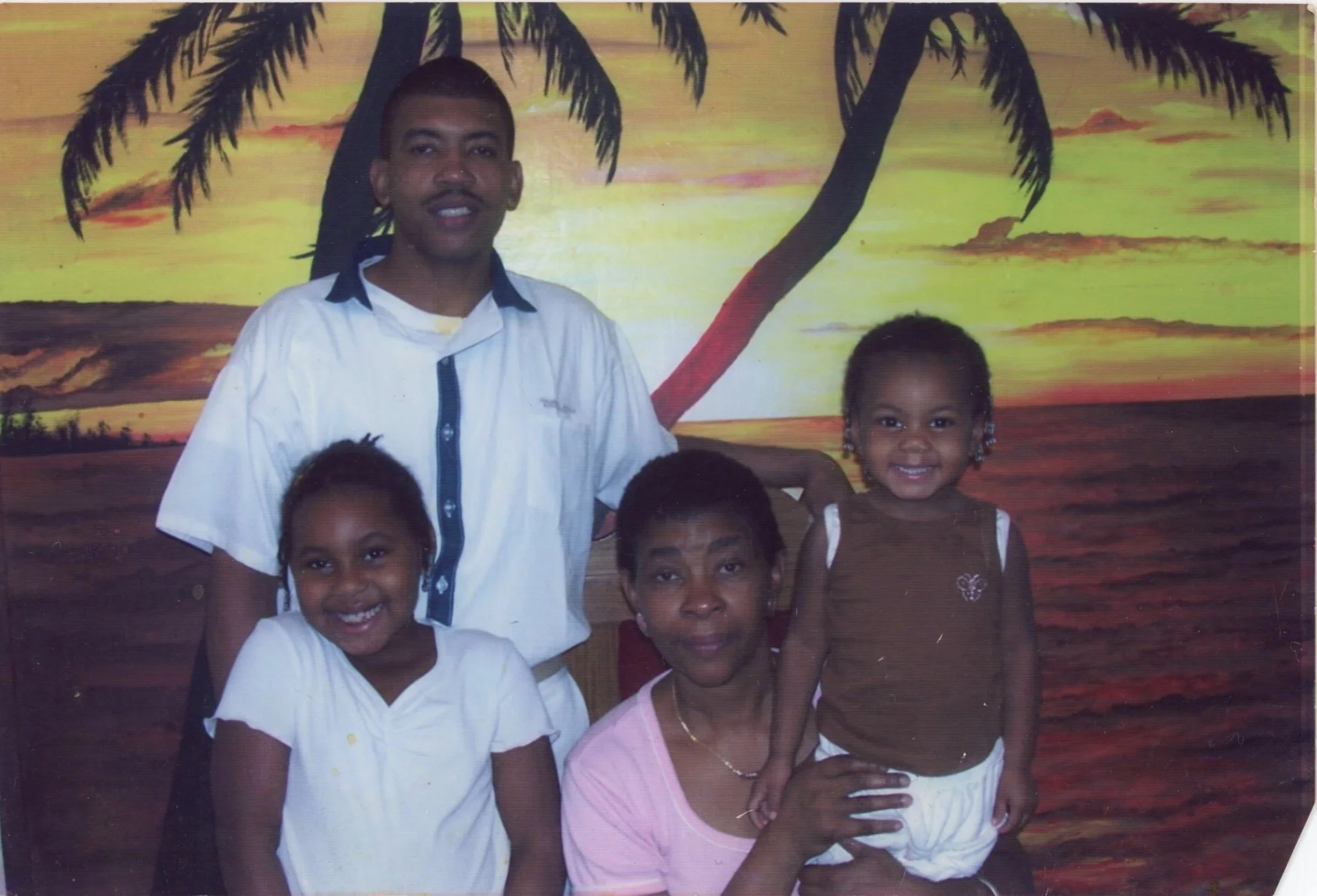 A family of five posing in front of a painted tropical sunset scene with palm trees.