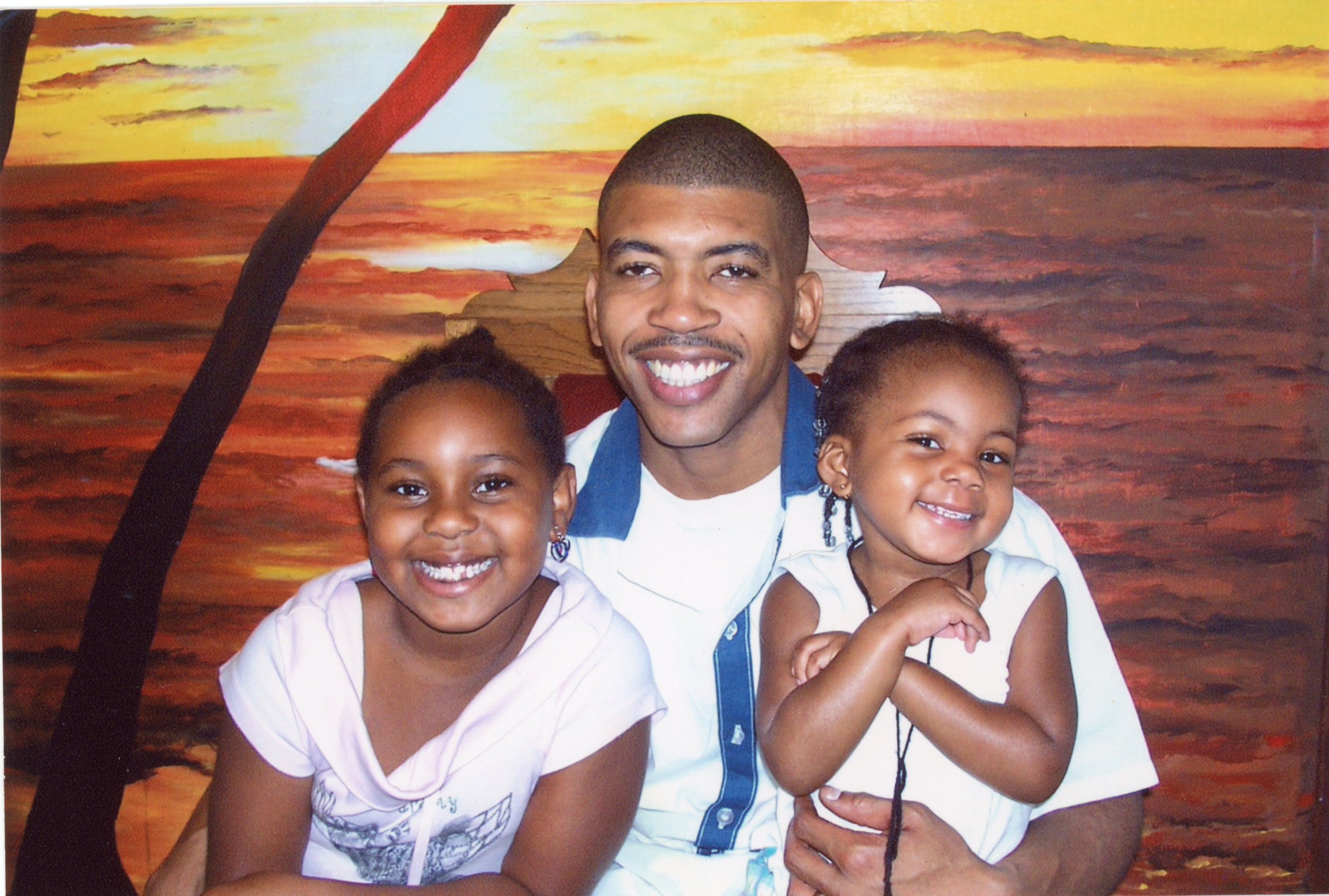 A man smiling while holding two young girls, with a painted sunset and ocean background.