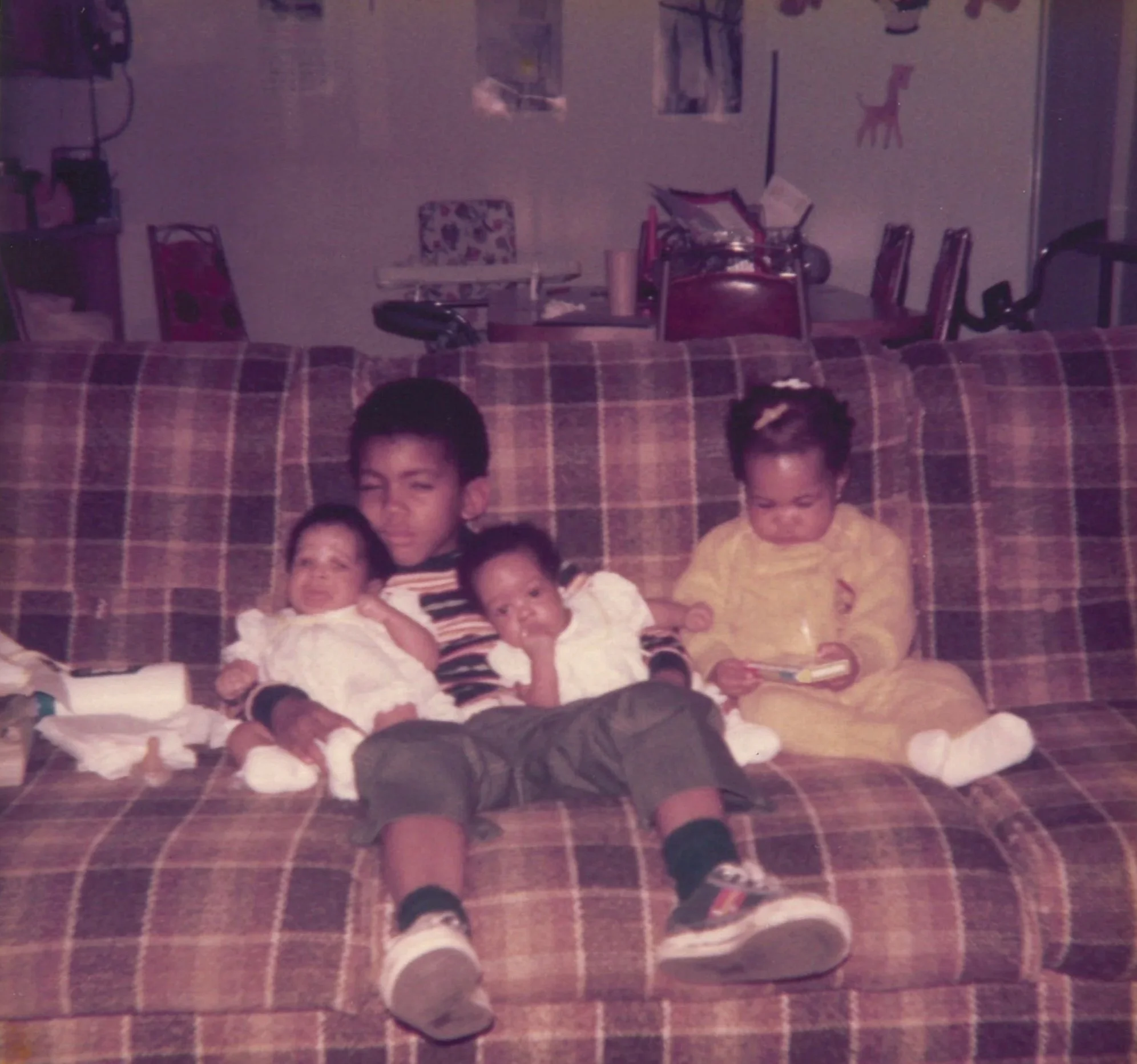 A young boy sitting on a plaid couch with three young children, two of whom are infants and one a toddler, in a cozy living room with children's artwork on the wall.