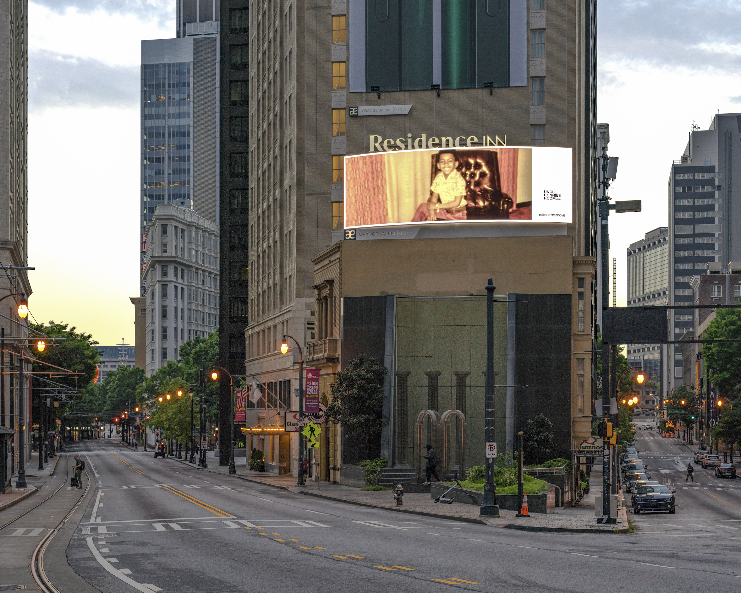 Empty city street with tall buildings, streetlights, and a digital billboard displaying an advertisement with a vintage photo of a child. The scene appears to be at dawn or dusk with minimal traffic.