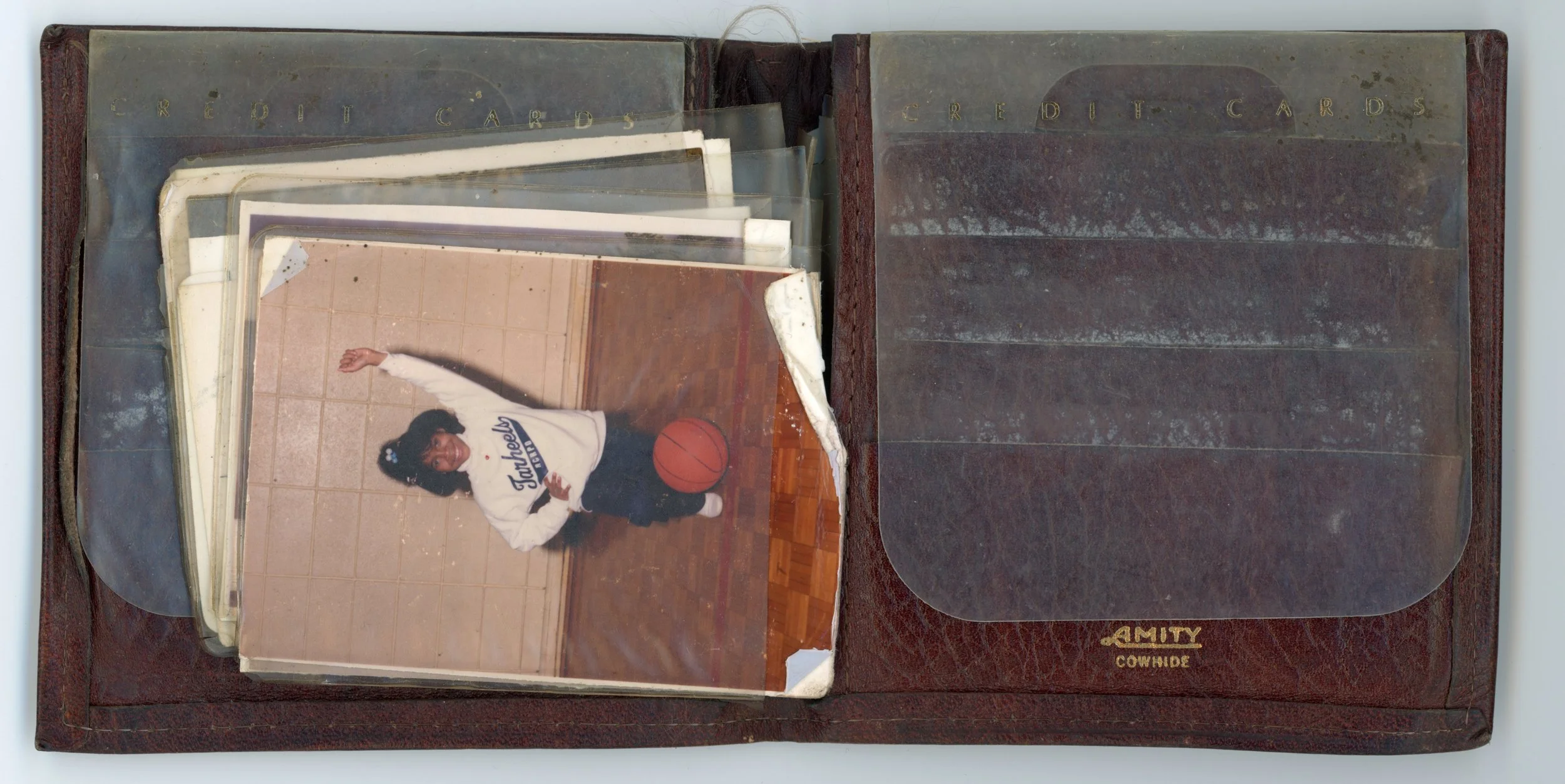 Open vintage photo album with plastic page protectors containing photographs and cards, featuring a young girl playing basketball indoors.