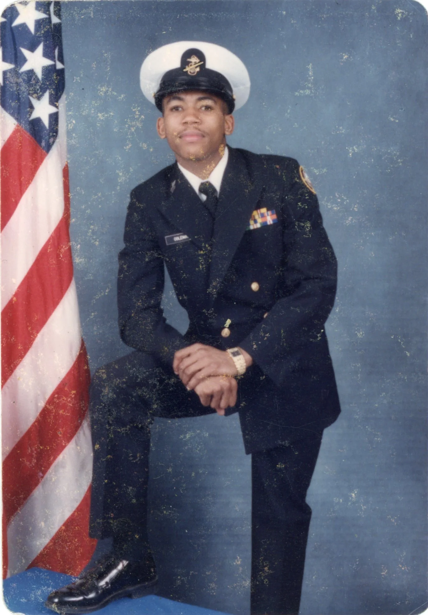 A young man in a United States military dress uniform, sitting with one foot on a stool and leaning forward slightly, poses next to an American flag against a blue background.