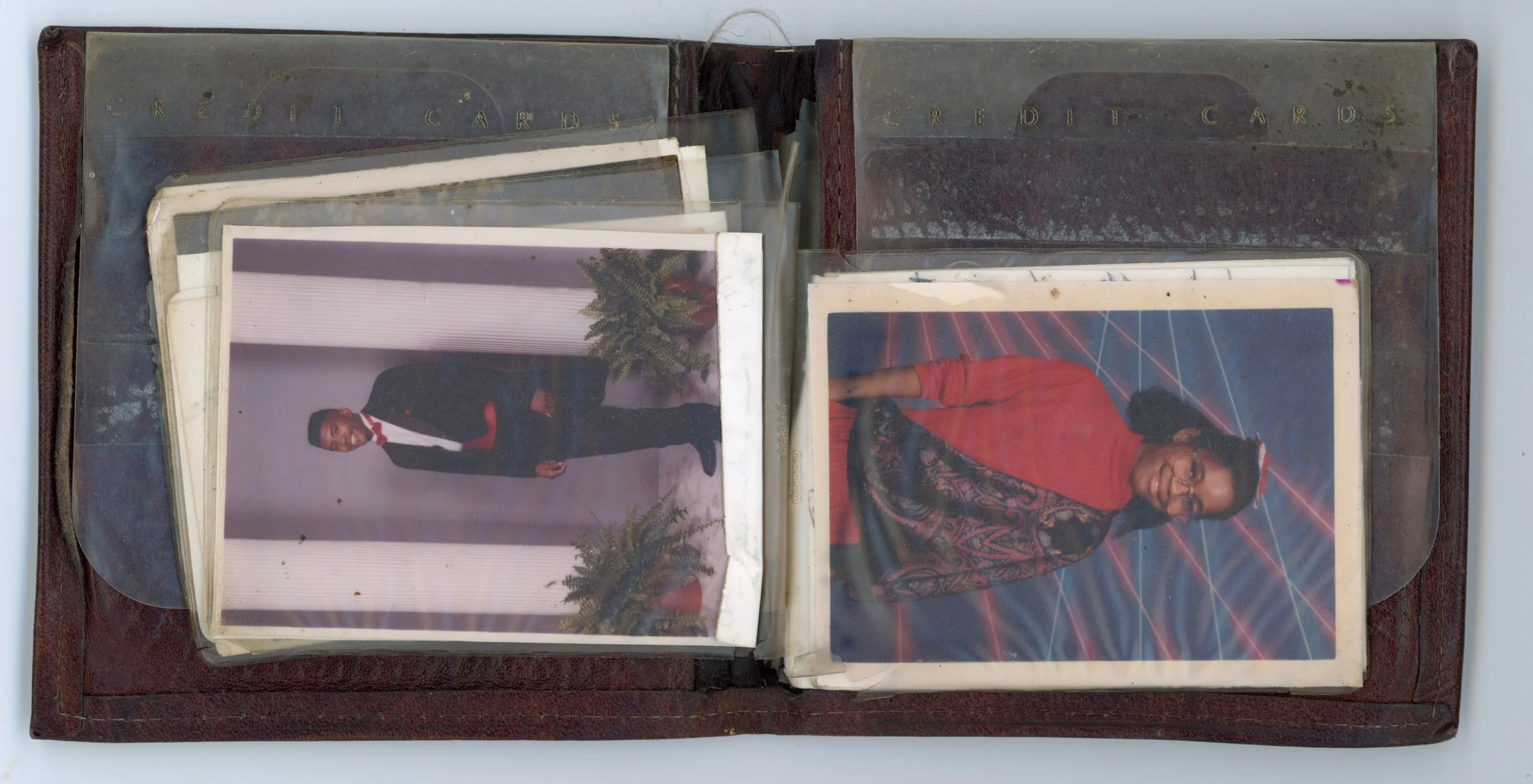 An open photo album with worn pages, containing three photographs of a woman and a man. The man is dressed in a tuxedo and pink bow tie, standing outdoors with plants and white columns behind him. The woman is dressed in a red outfit with a black pat