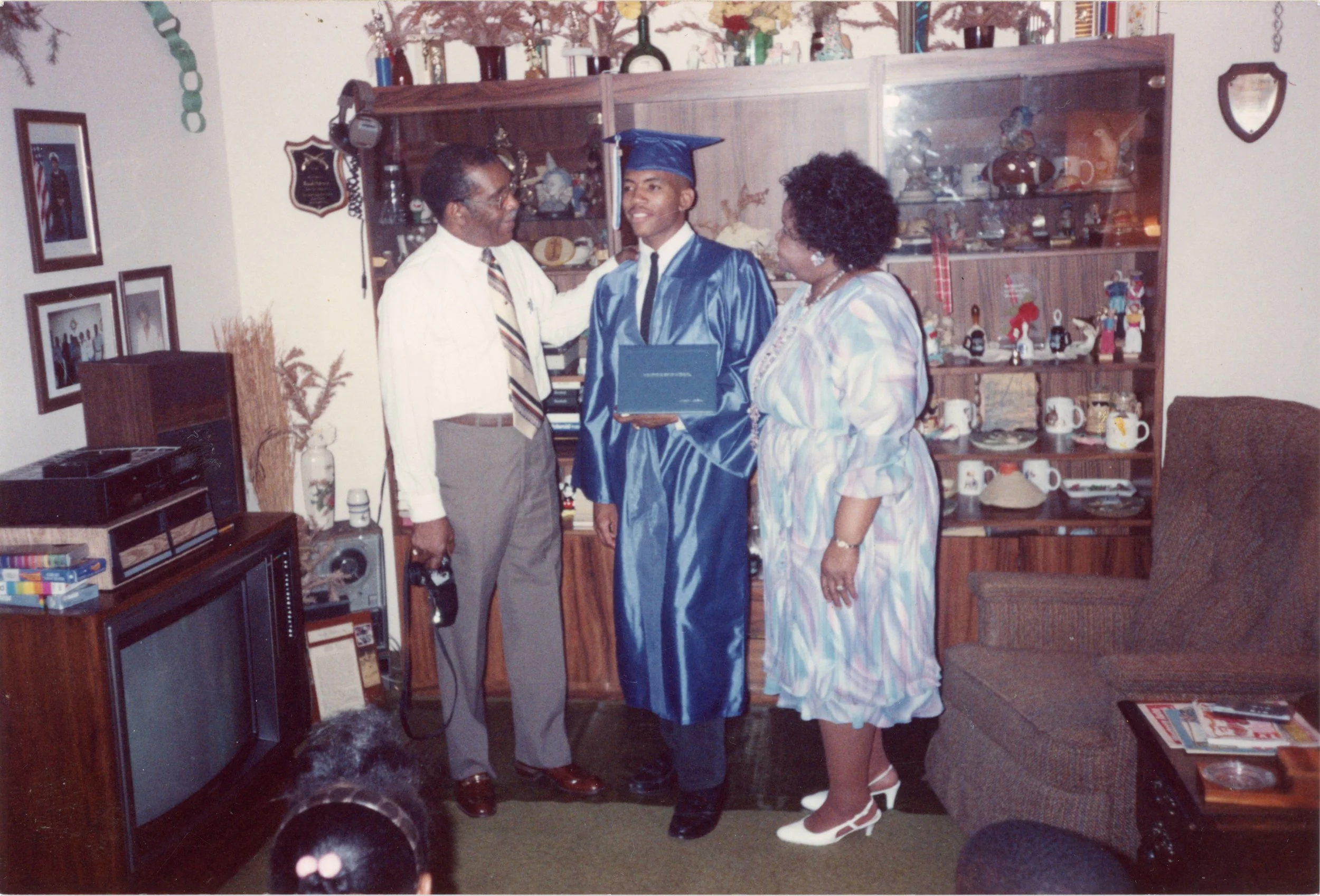 A young man in a blue graduation cap and gown holding a diploma, standing with his parents as he receives his diploma in a living room decorated with family photos, trophies, and knickknacks.