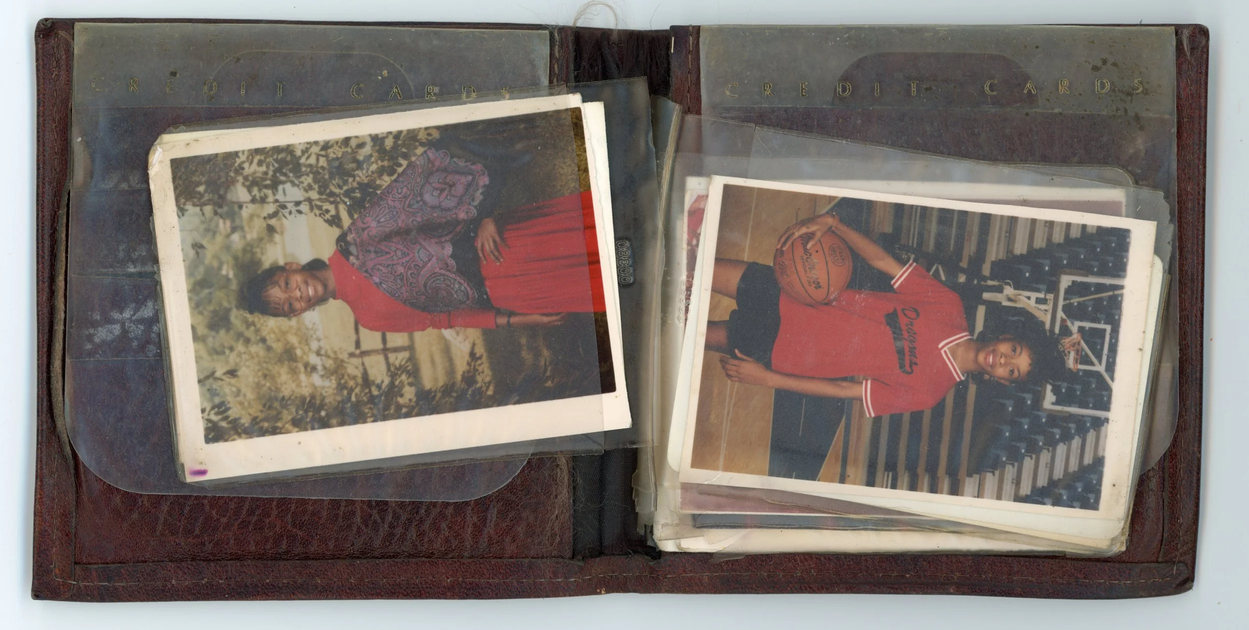 An open vintage photo album containing two photographs, one of a woman standing outdoors in a patterned scarf and red dress, and the other of a young woman in a red basketball uniform holding a ball in a gymnasium.
