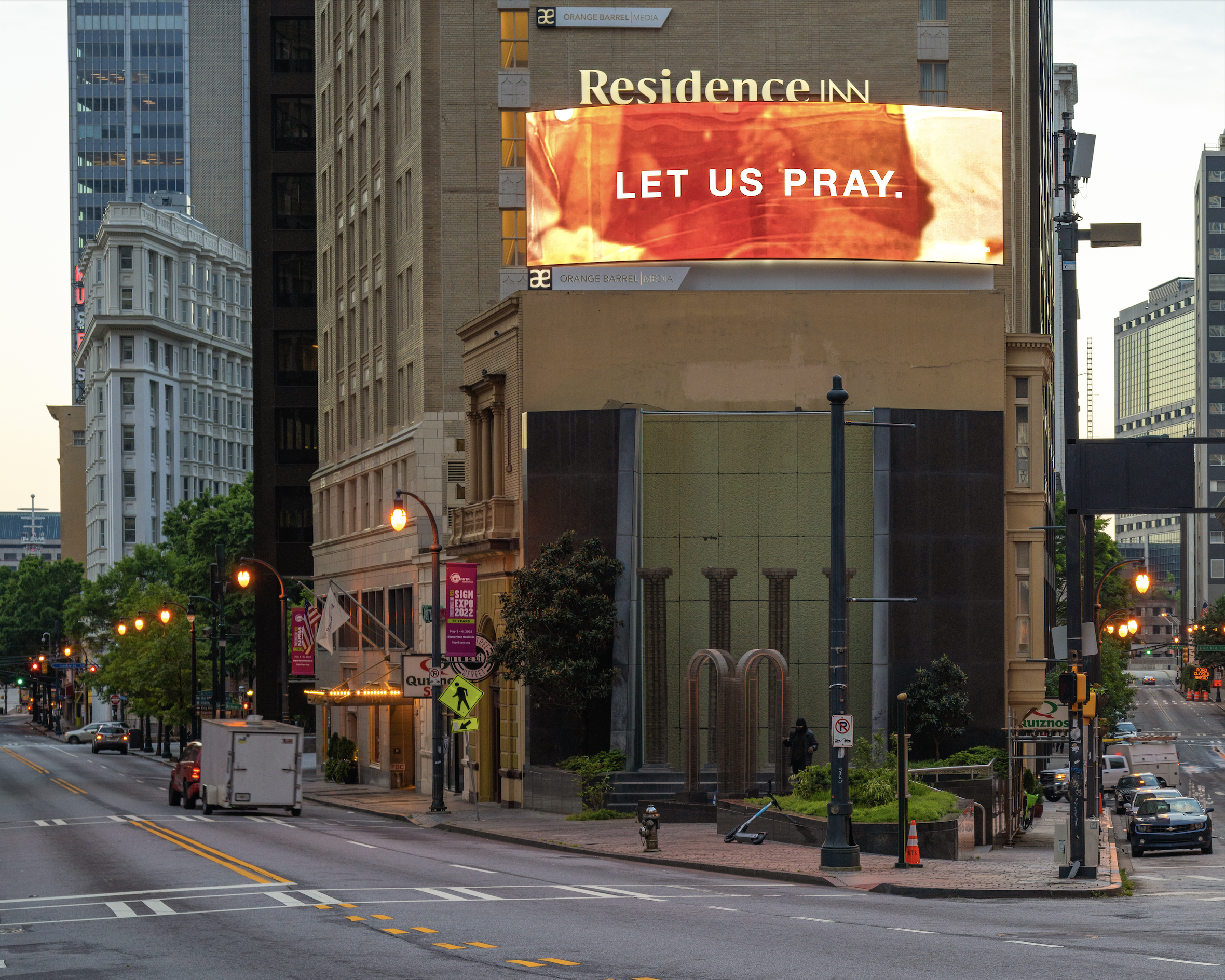City street intersection with buildings, street lamps, pedestrian crosswalk, and a digital billboard displaying the message "LET US PRAY." The area appears to be in downtown with glass and stone buildings, some trees, and a few parked and moving vehicles.