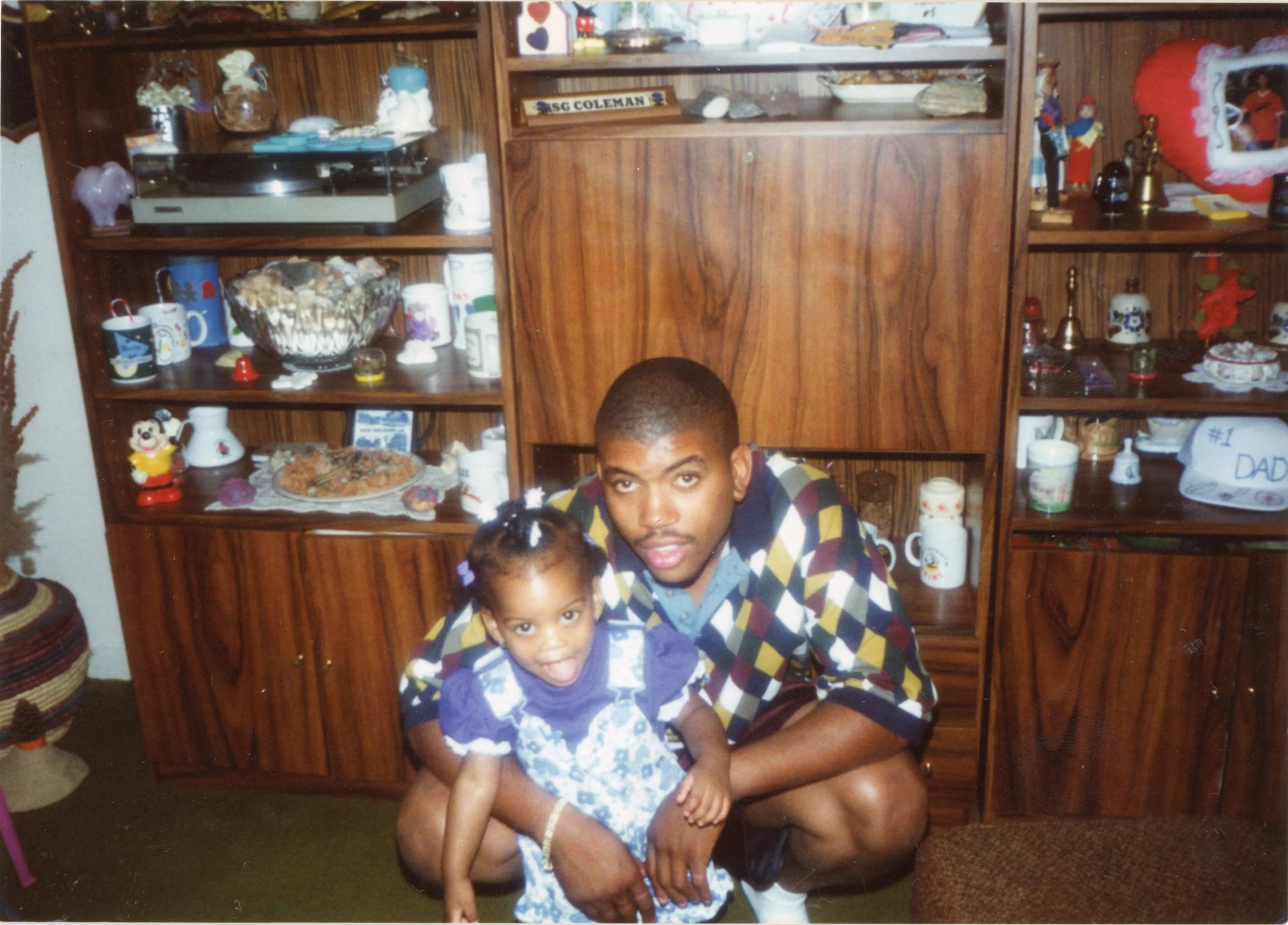 A young man kneeling next to a girl in a living room, with a wooden display cabinet filled with various holiday-themed decorations and mugs in the background.
