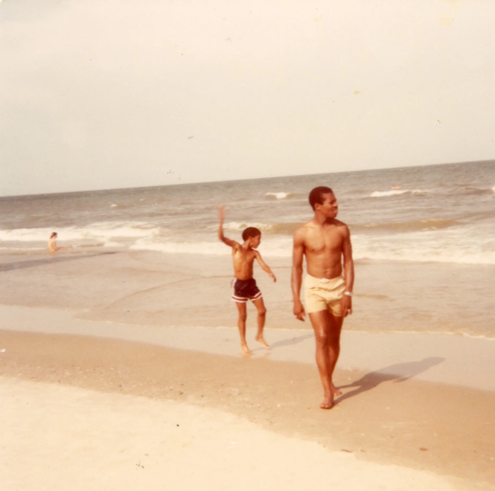 Two young boys walking along the sandy beach near the water, with one boy in the background playing near the shoreline and another boy closer to the camera walking barefoot.