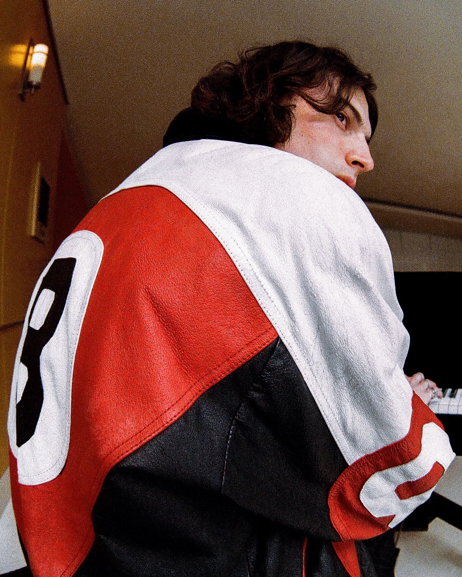 A person with brown wavy hair wearing a red, black, and white sports jacket, sitting in front of a keyboard.