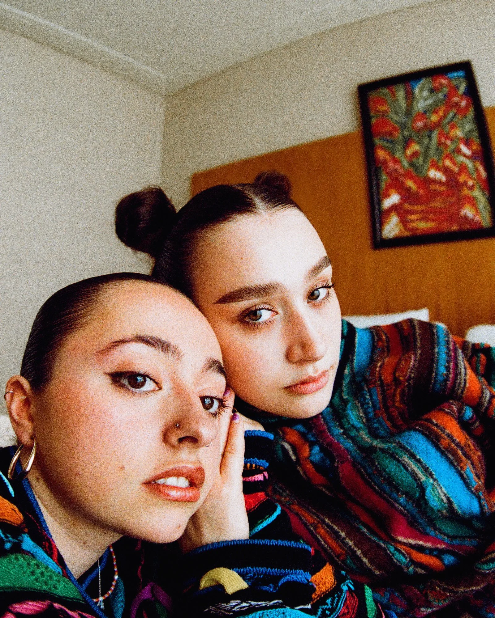 Two young women with dark hair and bold makeup, posing closely together indoors, with a colorful painting on the wall behind them.