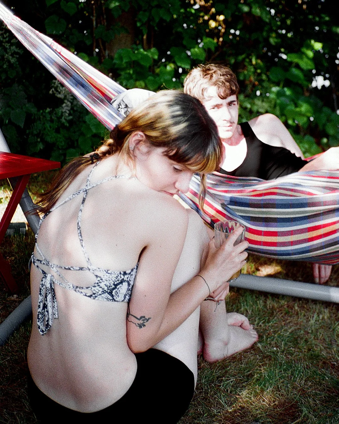A young woman with a snake print bikini top sitting on the grass, looking at her phone, with a young man laying in a colorful hammock behind her.