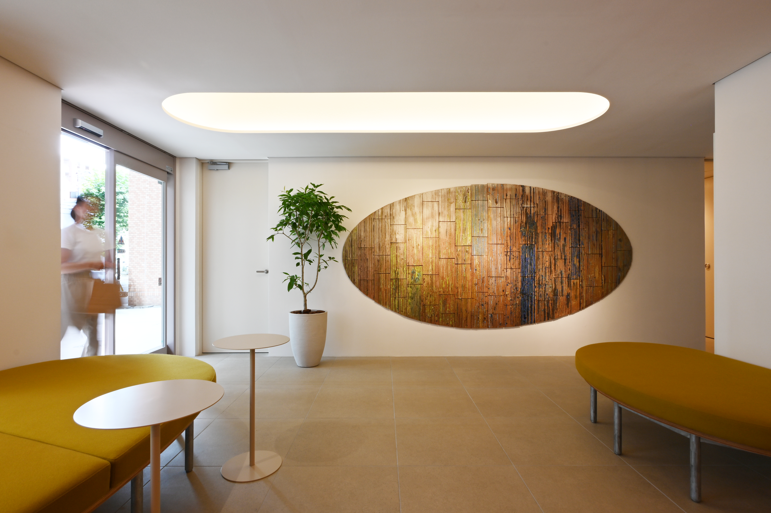 Modern interior space with yellow cushioned seating, small white round tables, a tall potted plant, and a large wooden wall art piece on white wall. Large windows and overhead lighting provide natural and artificial light.