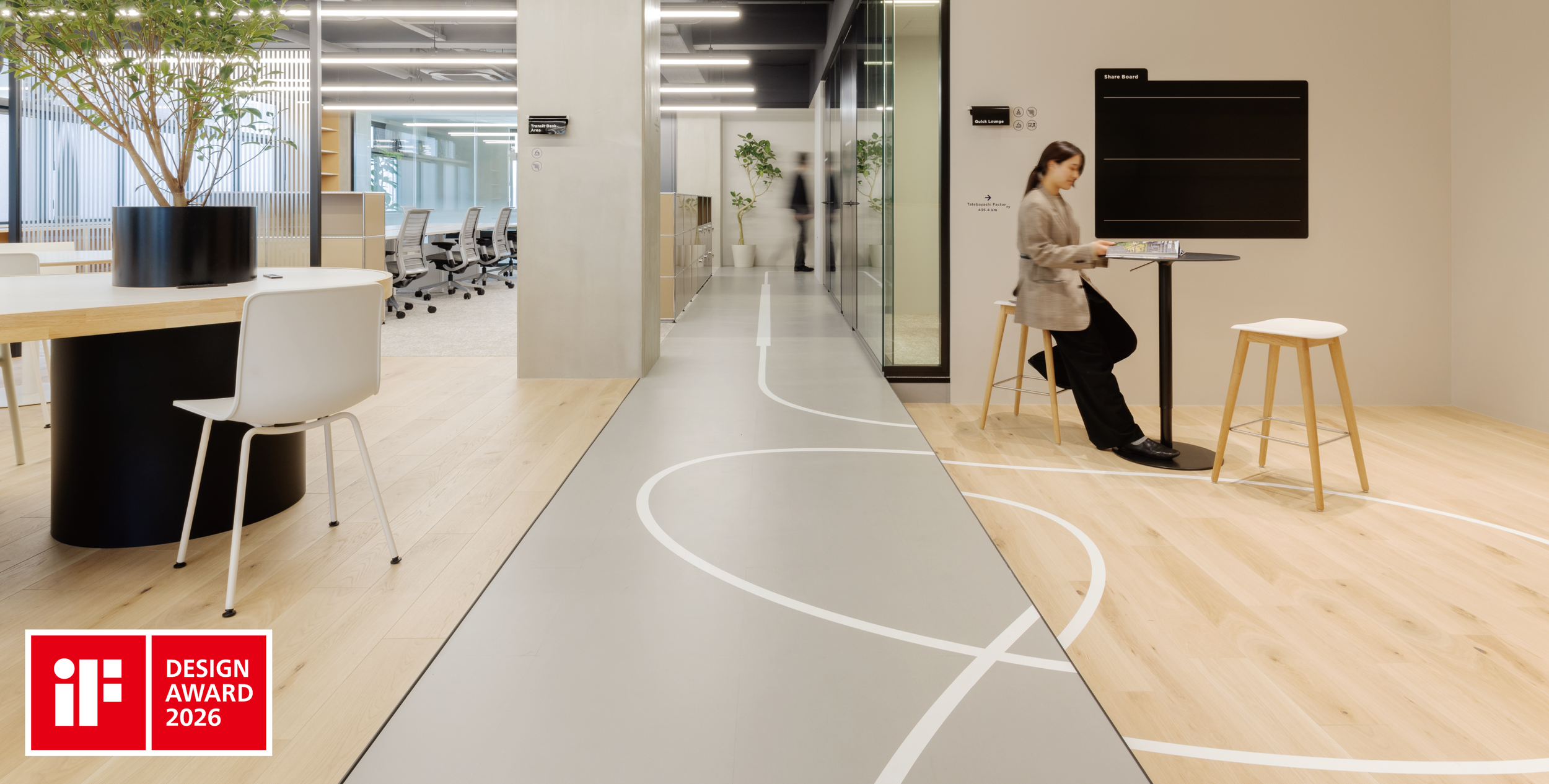 Modern office interior with a winding line pattern on the floor, a woman sitting at a small table reading a magazine, and a section with conference rooms and workstations in the background. There is an iF Design Award 2026 logo in the bottom left corner.