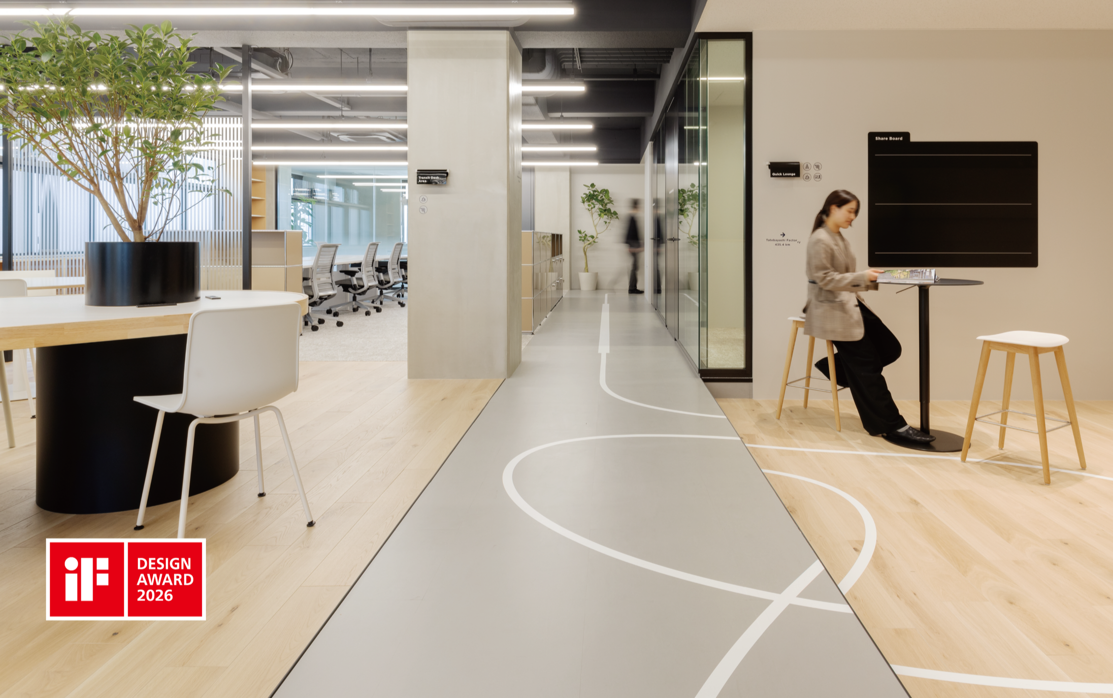 Modern office interior with a winding line pattern on the floor, a woman sitting at a small table reading a magazine, and a section with conference rooms and workstations in the background. There is an iF Design Award 2026 logo in the bottom left corner.