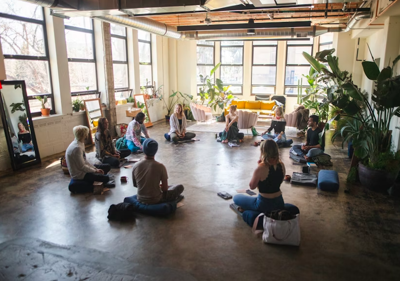 A group of people sitting in a circle on the floor in a bright room with large windows, plants, and a yellow couch, participating in a group meditation or yoga session.