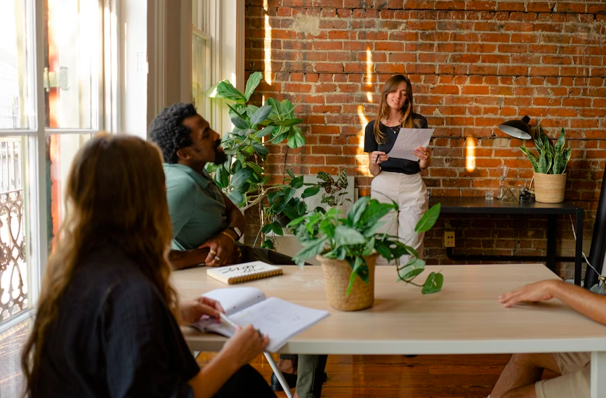 A woman stands in front of a brick wall holding papers, speaking to a group during a meeting in a bright room with large windows and green plants.