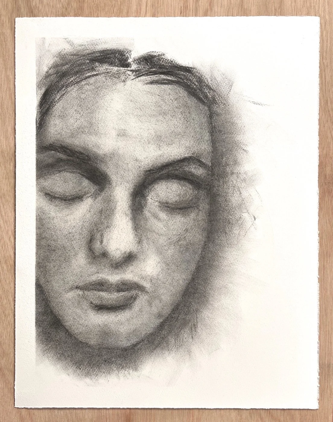 Full sheet view of a charcoal drawing depicting a face with closed eyes, surrounded by large areas of untouched paper.